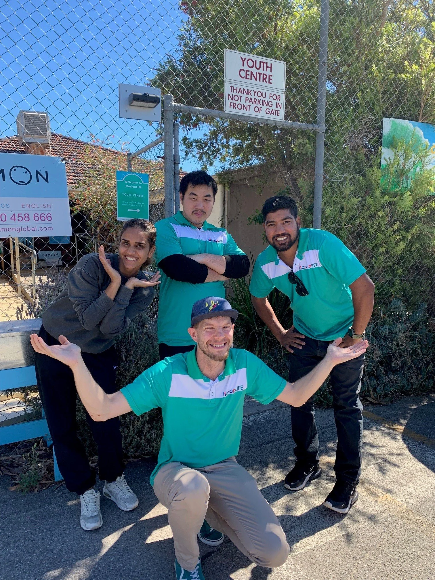 Group of five smiling young people posing outdoors in front of a chain-link fence with signs, one reading 'YOUTH CENTRE' and another with a slogan, under a clear blue sky.