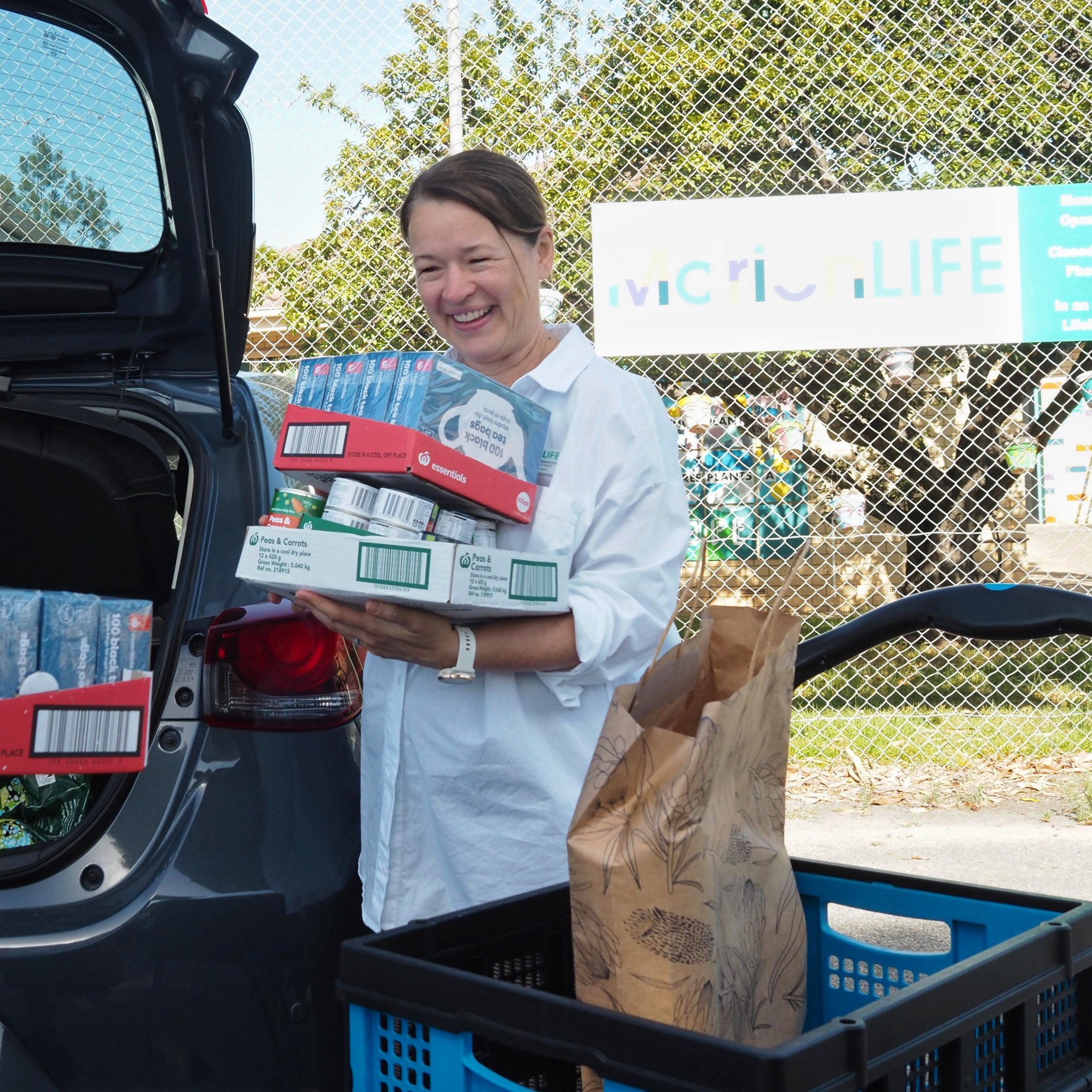 A woman loading or unloading grocery items, including canned goods and boxed food, from the trunk of a car. The scene is outdoors with a chain-link fence and trees in the background.