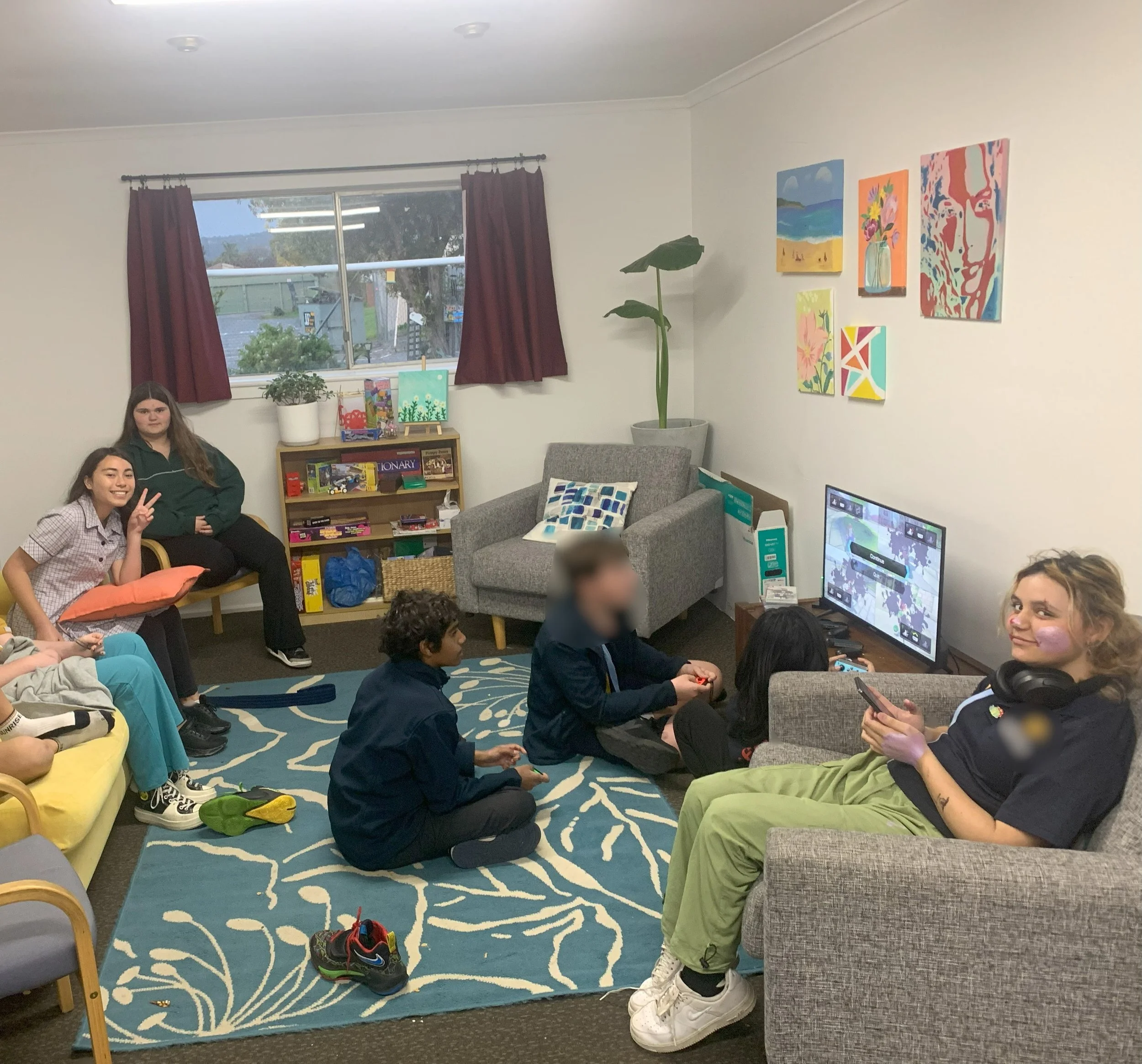 Group of people gathered in a cozy living room with colorful paintings on the wall, a window with red curtains, a gray armchair, a bookshelf, and a television, engaged in conversation and activities.