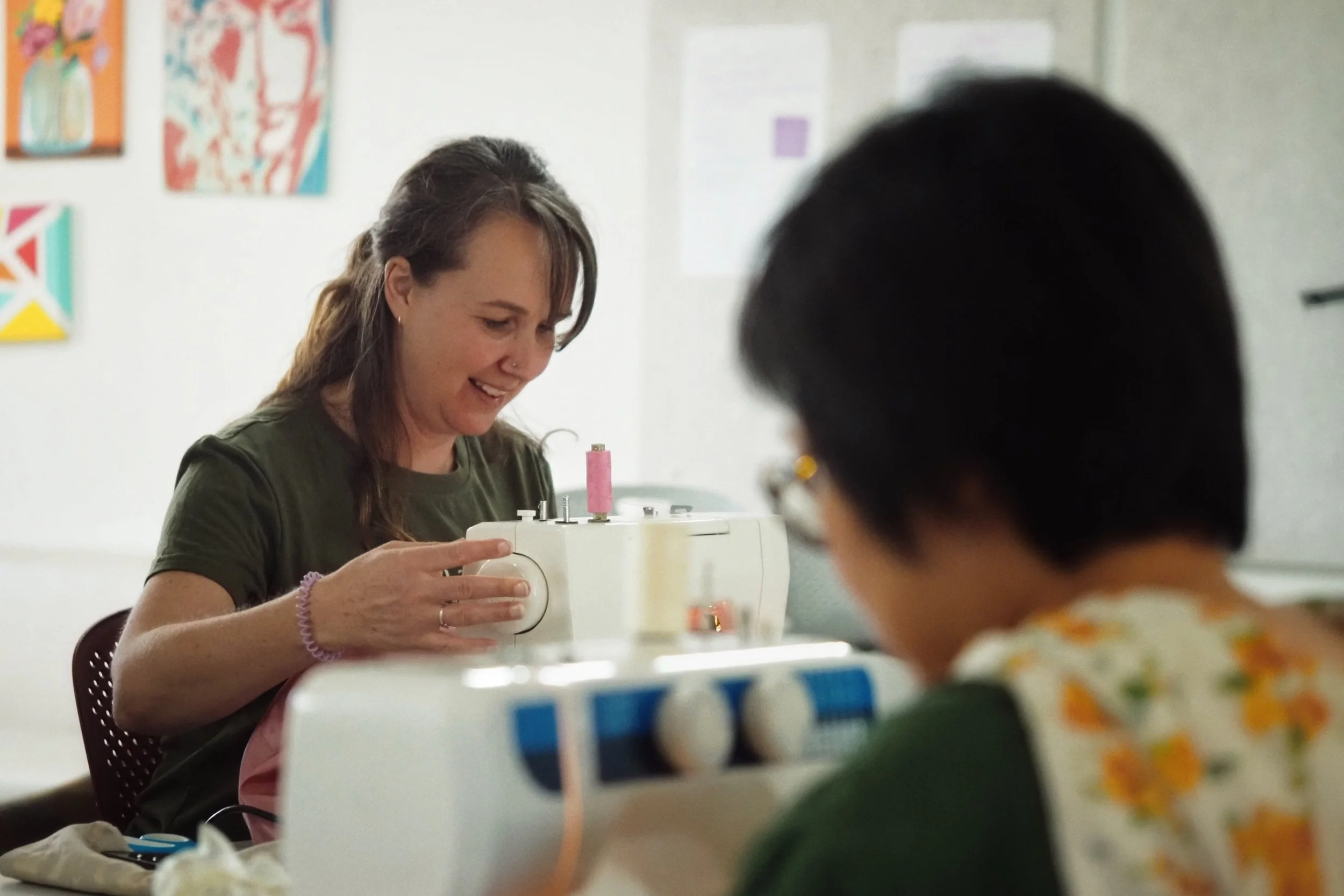 A woman sewing on a white sewing machine with a young child in the foreground, blurred. The woman is smiling, wearing a dark green shirt, and has long brown hair.