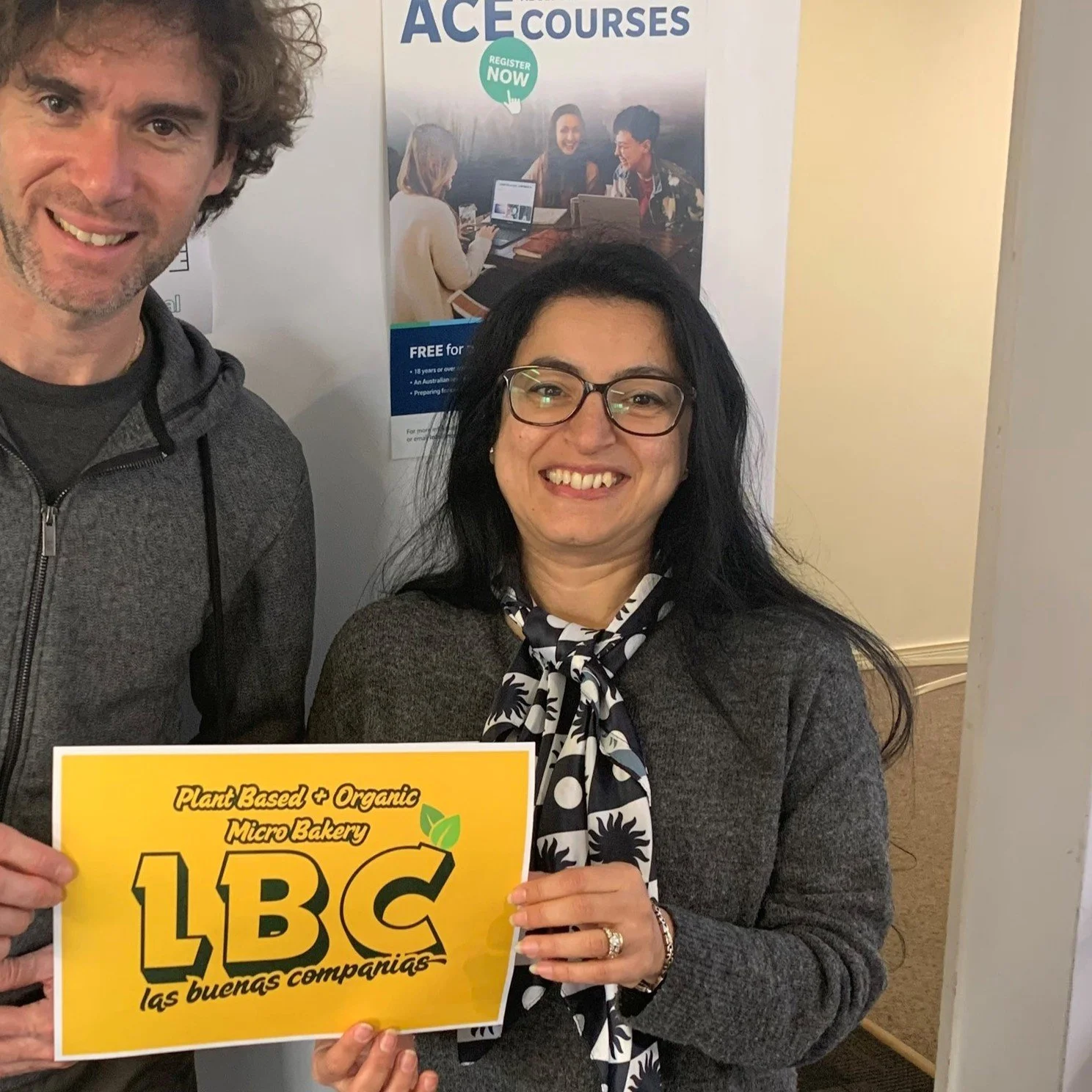 A woman with black hair, glasses, and a patterned scarf, smiling and holding a yellow sign that says "LBC Las buenas compañías." Partially visible man with curly hair standing next to her. Behind them, a poster with the words "ACE COURSES" and a photo of three people sitting at a table with laptops, with a green "Register Now" button.