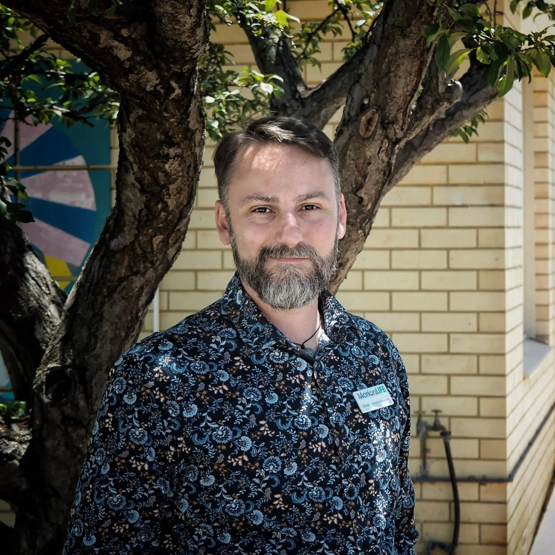 A man with a beard and short hair standing outdoors next to a tree, wearing a dark blue floral shirt and a name tag that says 'MarionLIFE,' with a yellow brick wall in the background.