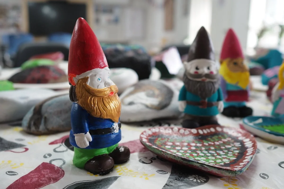 Colorful ceramic gnome figurines, including a gnome with a red hat, blue shirt, and green pants, on a decorated table with rocks and other craft items in the background.