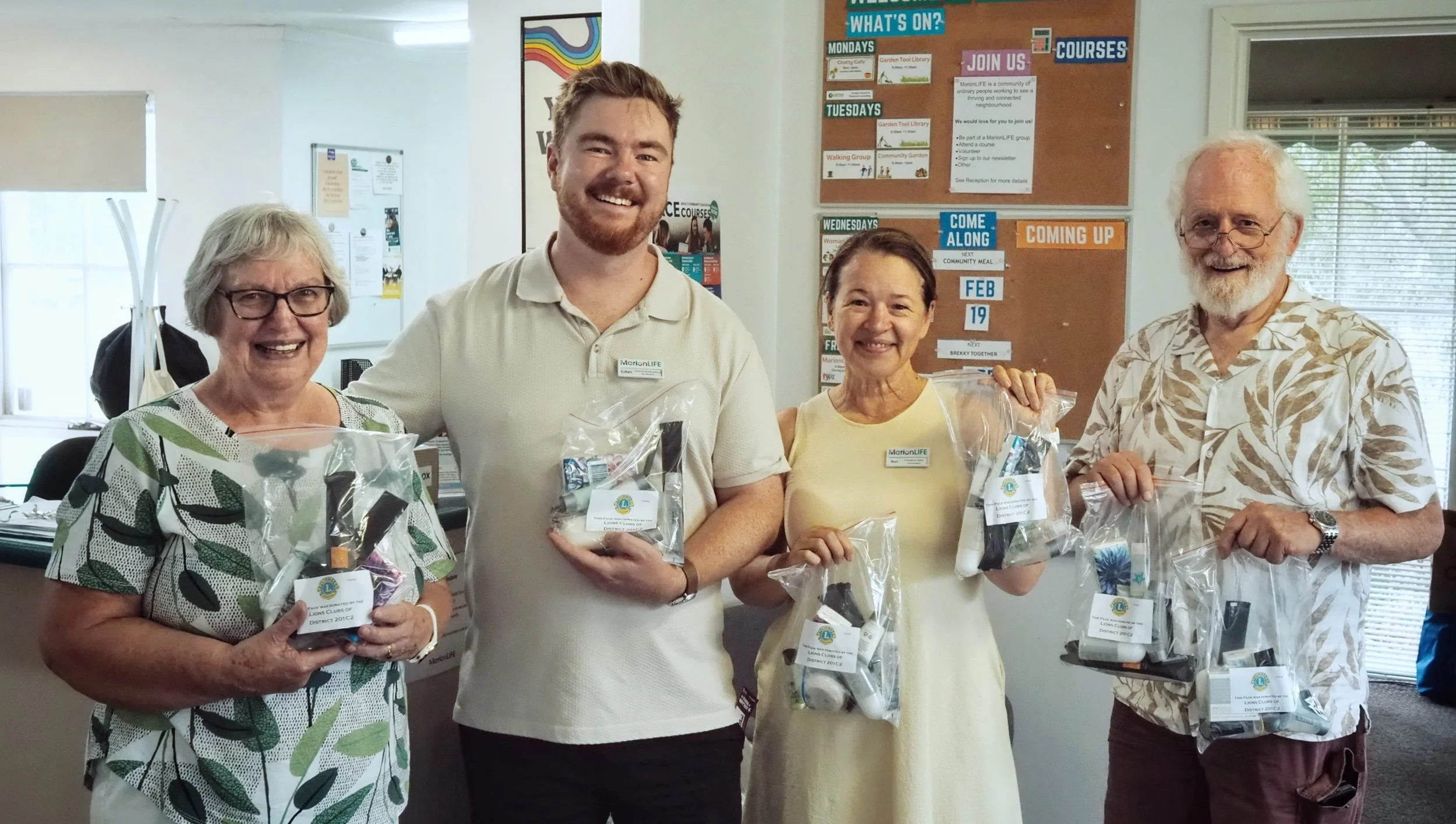 Four smiling people holding gift bags, standing in an office or community center with a bulletin board behind them. The bulletin board contains various notices and schedules.