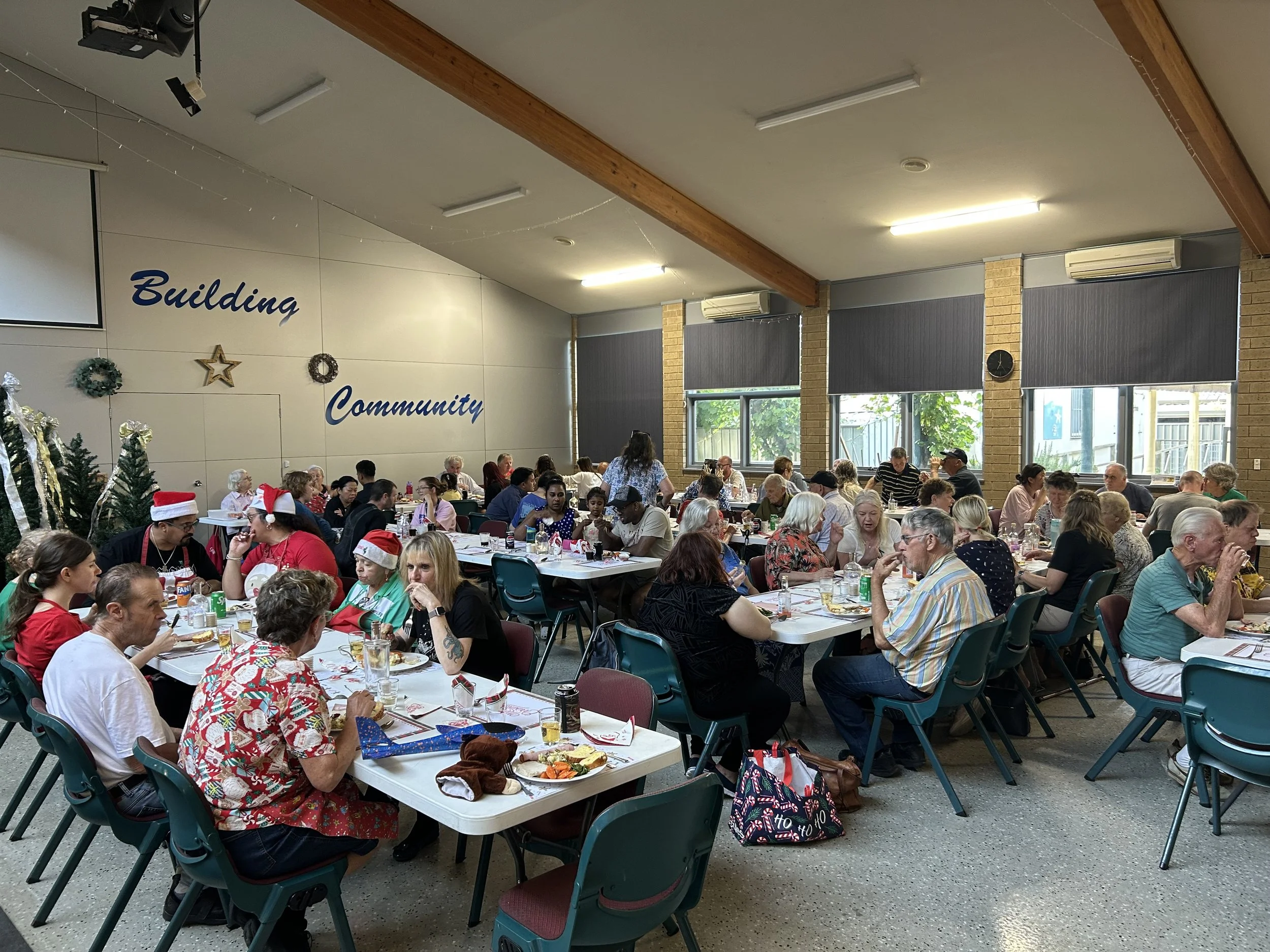 Community building event with people sitting at long tables, Christmas decorations, trees, and holiday attire, including Santa hats, inside a decorated hall.