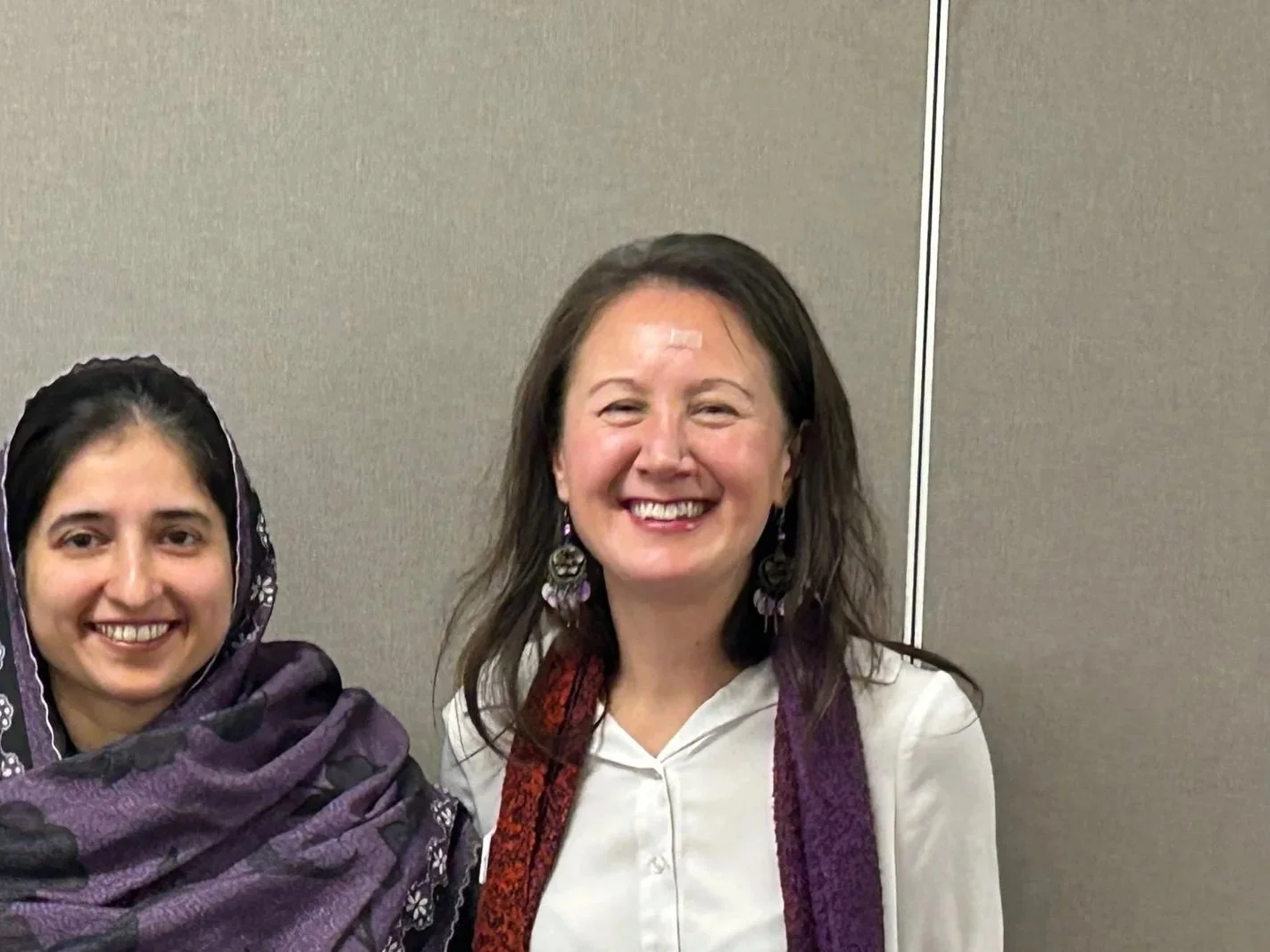 Two women smiling, standing against a plain beige wall. One woman wears a patterned shawl over her head and shoulders, the other wears large earrings and a purple scarf.