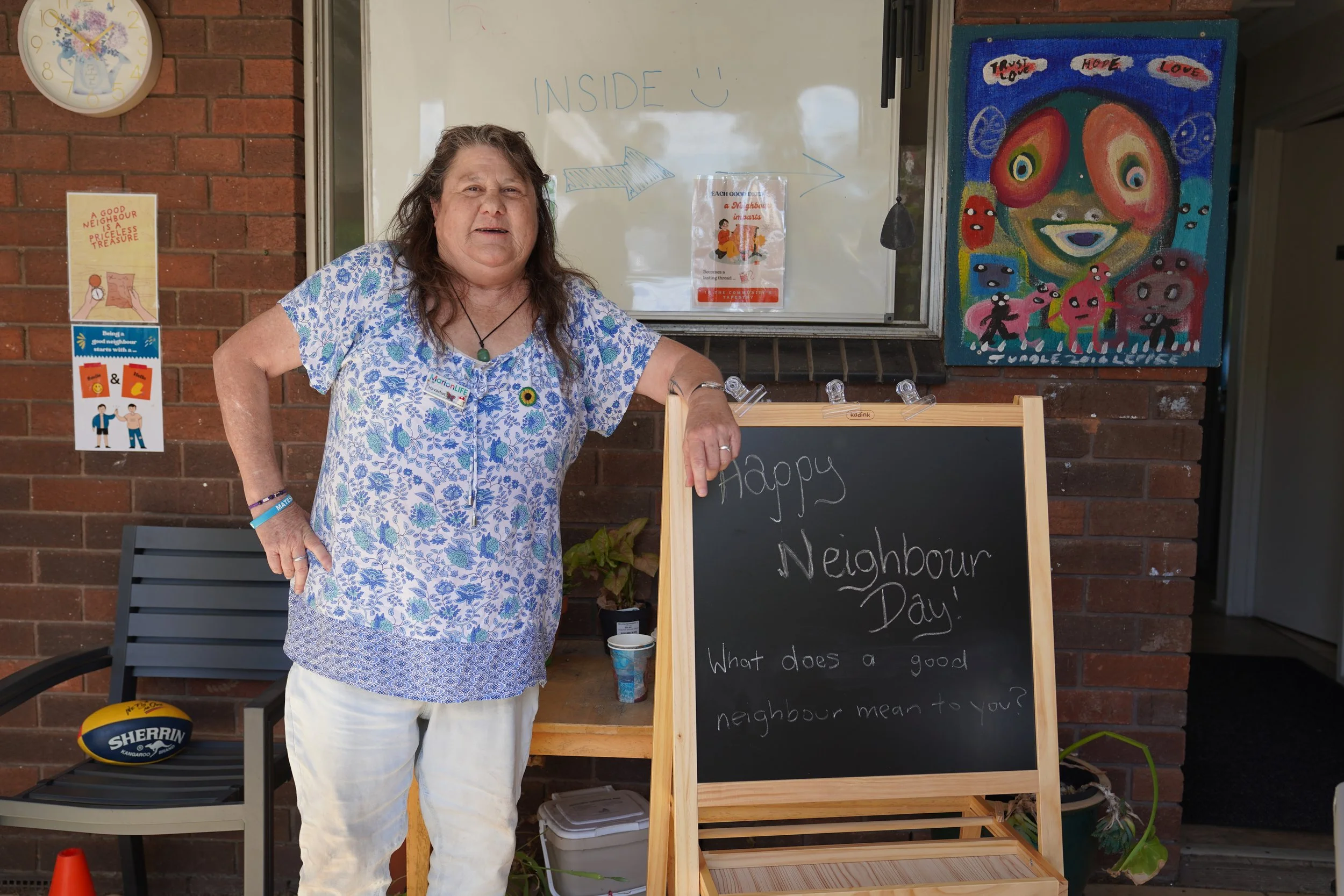 A woman with long, wavy brown hair wearing a blue and white floral blouse and white pants stands outside near a brick wall, smiling. Behind her is a whiteboard with the word 'INSIDE' and a smiley face drawn, and a colorful abstract art piece on the wall. To her right is a blackboard sign that reads 'Happy Neighbour Day!' with partial text below. On her left, there are various signs and a sports ball on a chair.