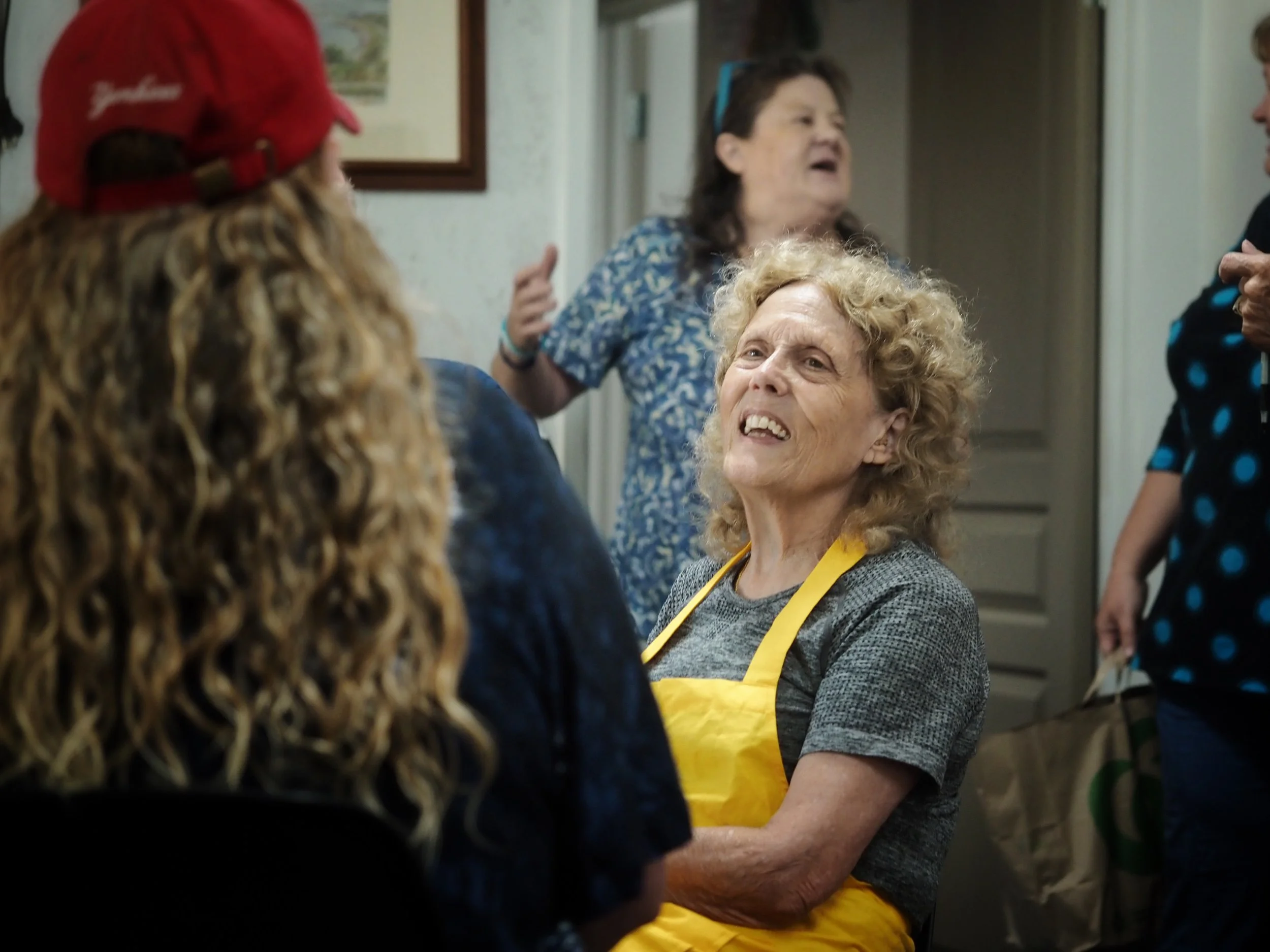 A woman with short brown hair wearing a gray shirt and yellow apron is speaking passionately with her eyes closed and mouth open, while another person with long curly hair and a red hat listens with their back to the camera in a room with framed pict