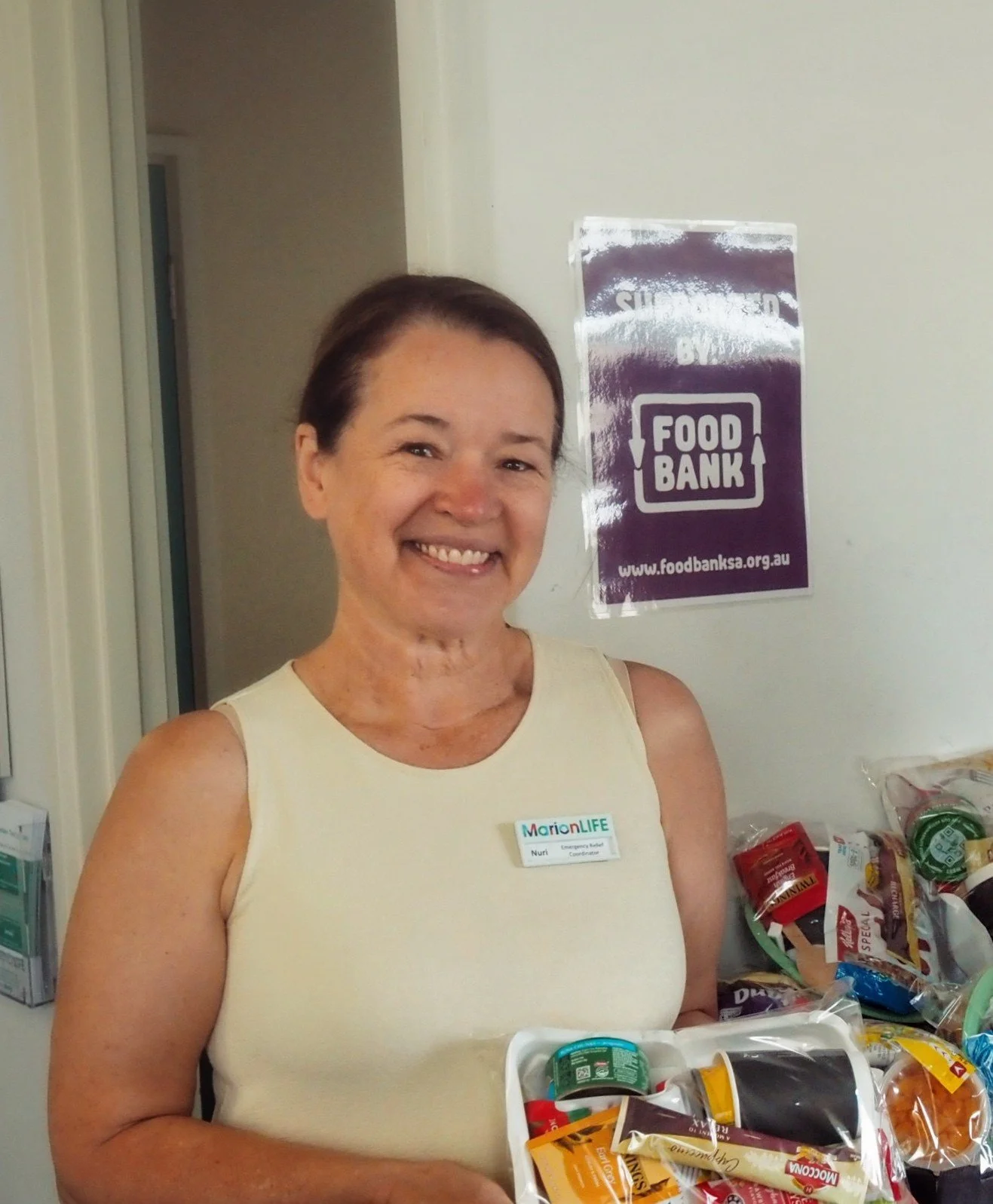 A woman with short dark hair and a beige sleeveless top smiling in front of a food bank sign and standing with food bags.