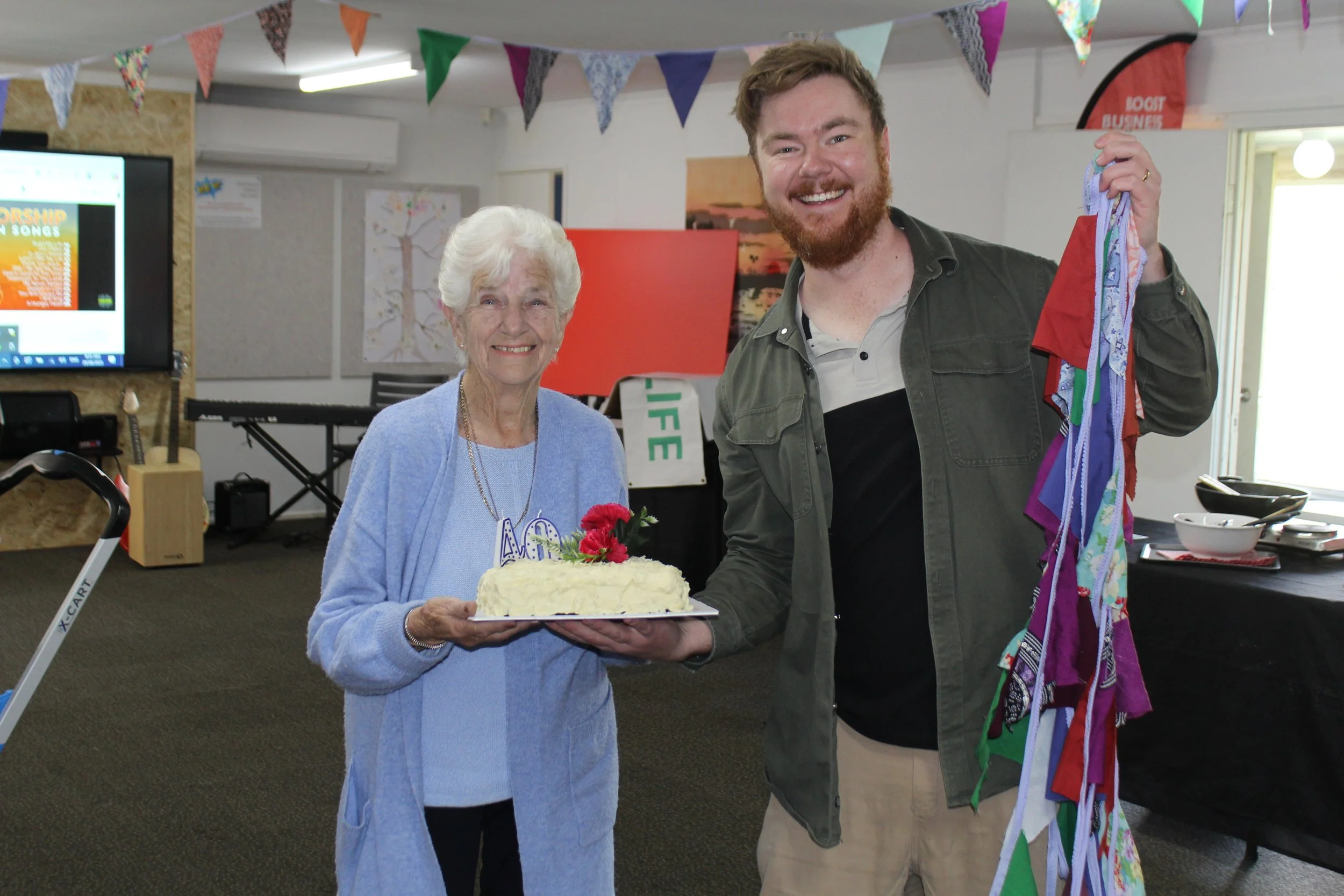An elderly woman and a young man celebrating together at a birthday party in an indoor setting decorated with colorful bunting. The elderly woman is holding a birthday cake with red flowers and numbers on top, while the young man, smiling, is holding