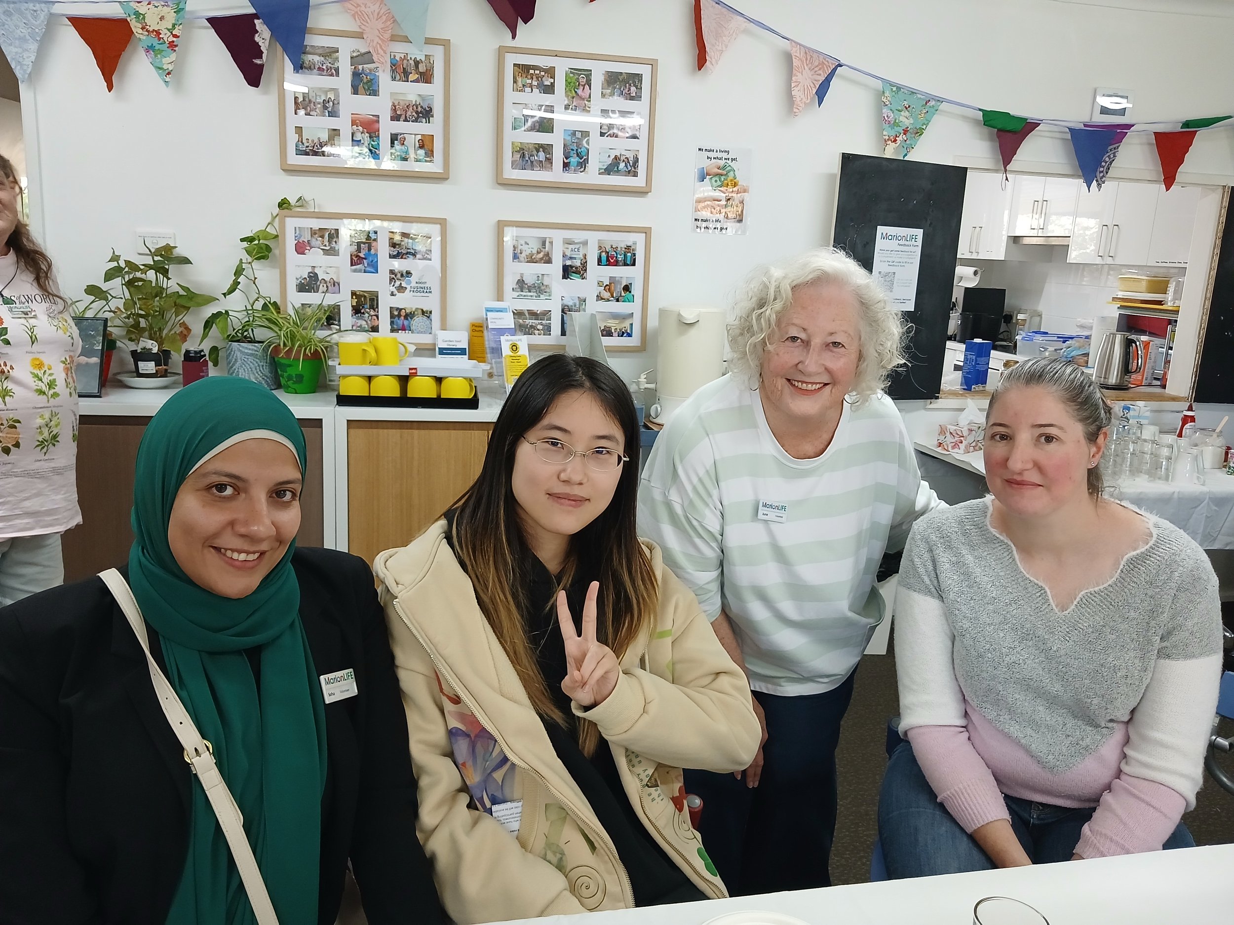 Group of five women and one man smiling and posing for a photo at an indoor event, with colorful bunting hanging overhead and framed photos on the wall in the background.