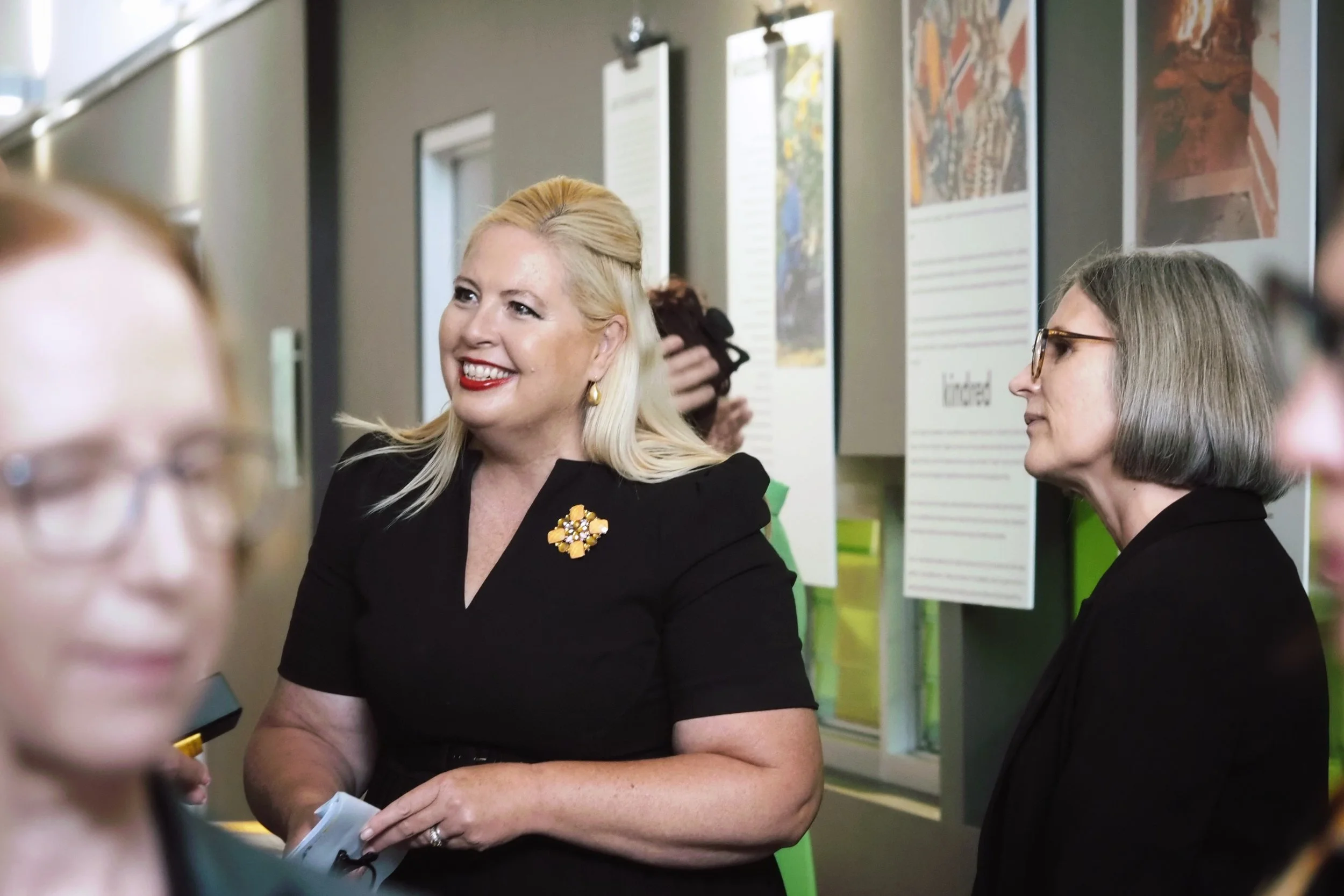 A woman with blonde hair, wearing a black dress with a yellow floral brooch, smiling while interacting with another woman, in an indoor setting with informational posters on the wall.