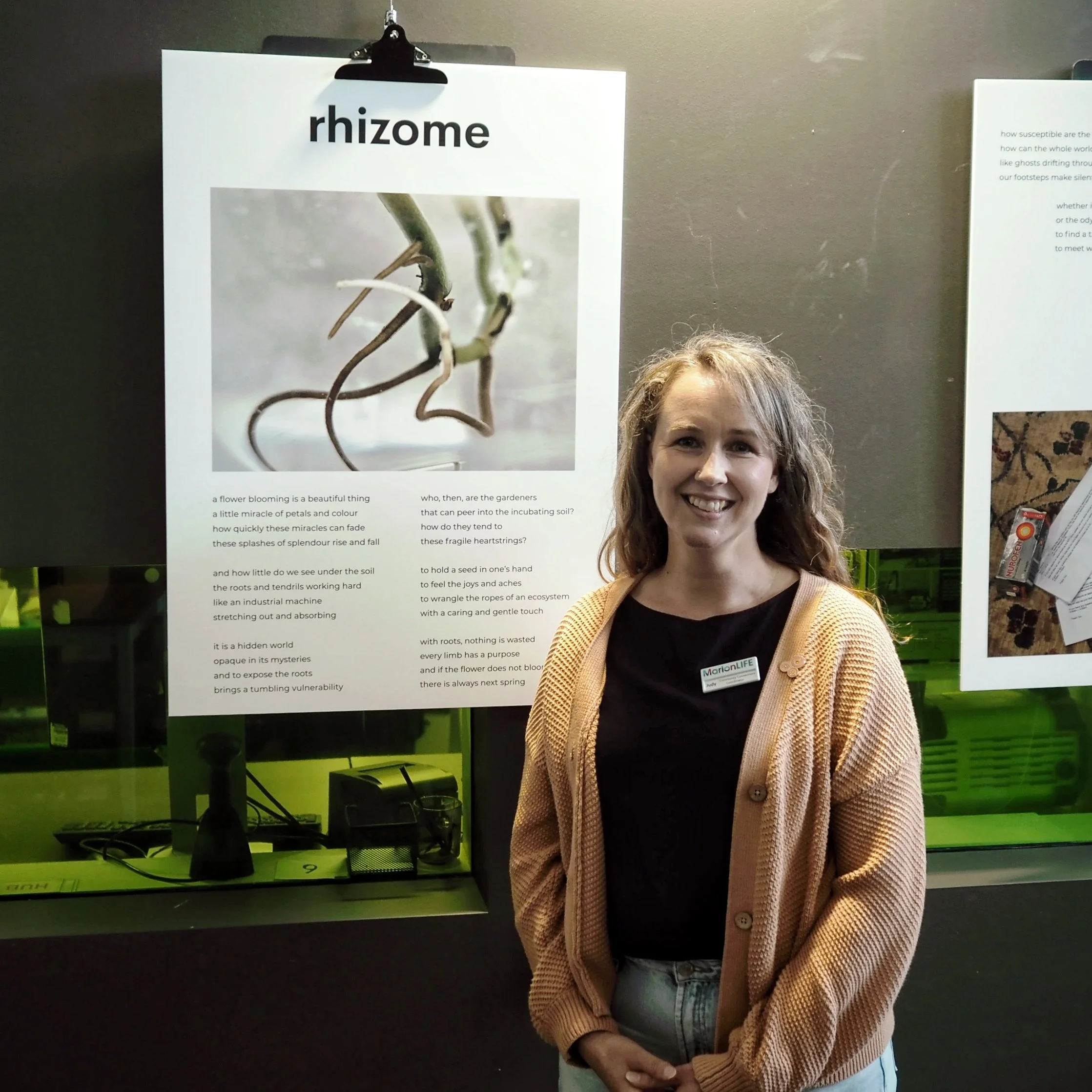 Young woman with curly hair smiling, wearing a beige cardigan and a badge, standing in front of display boards at an exhibit about rhizomes and plant roots.
