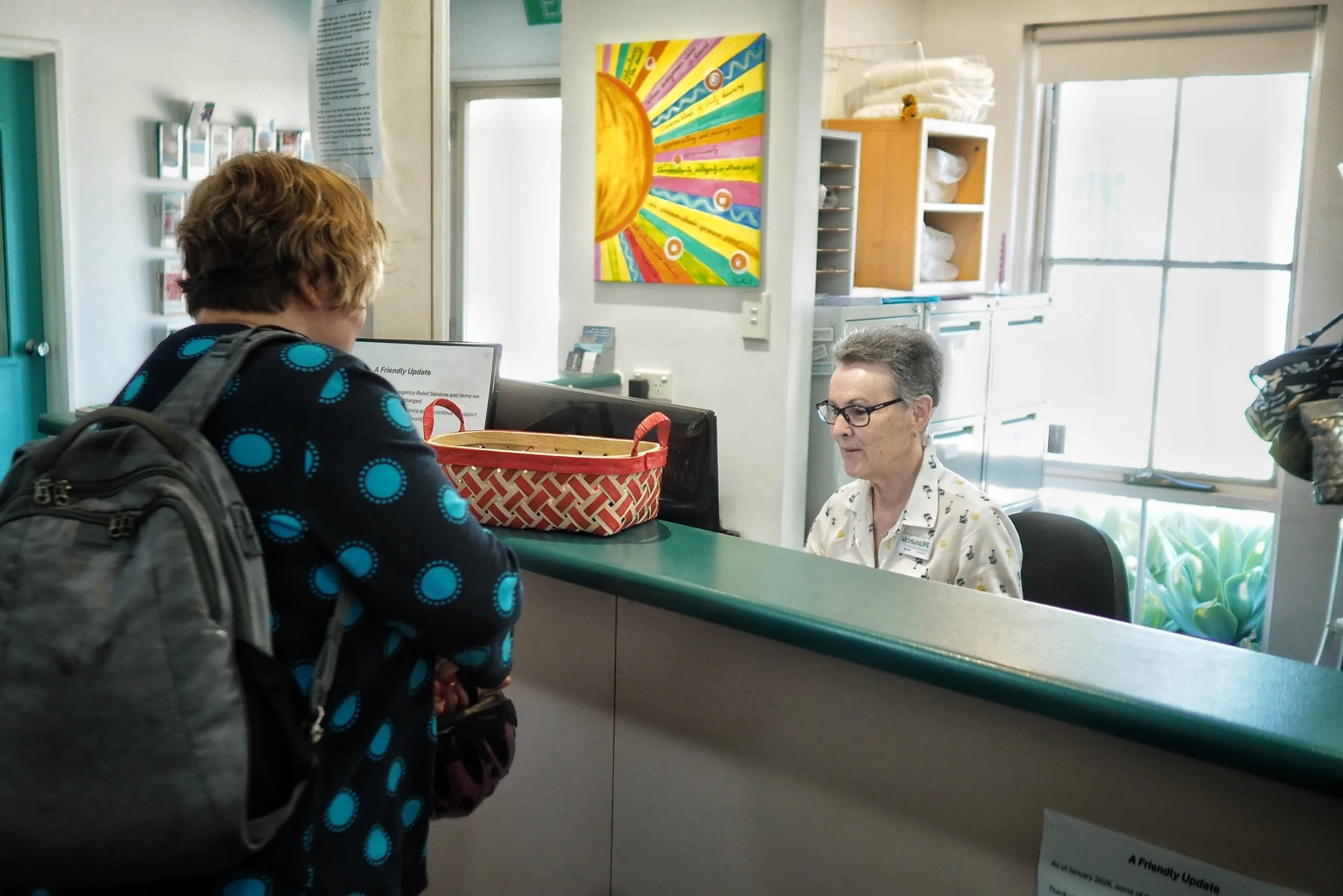 A person with curly red hair and a backpack interacting with a healthcare worker at a reception desk in a clinic or medical office. The doctor is an older woman with short gray hair and glasses, wearing a white shirt with small black patterns.
