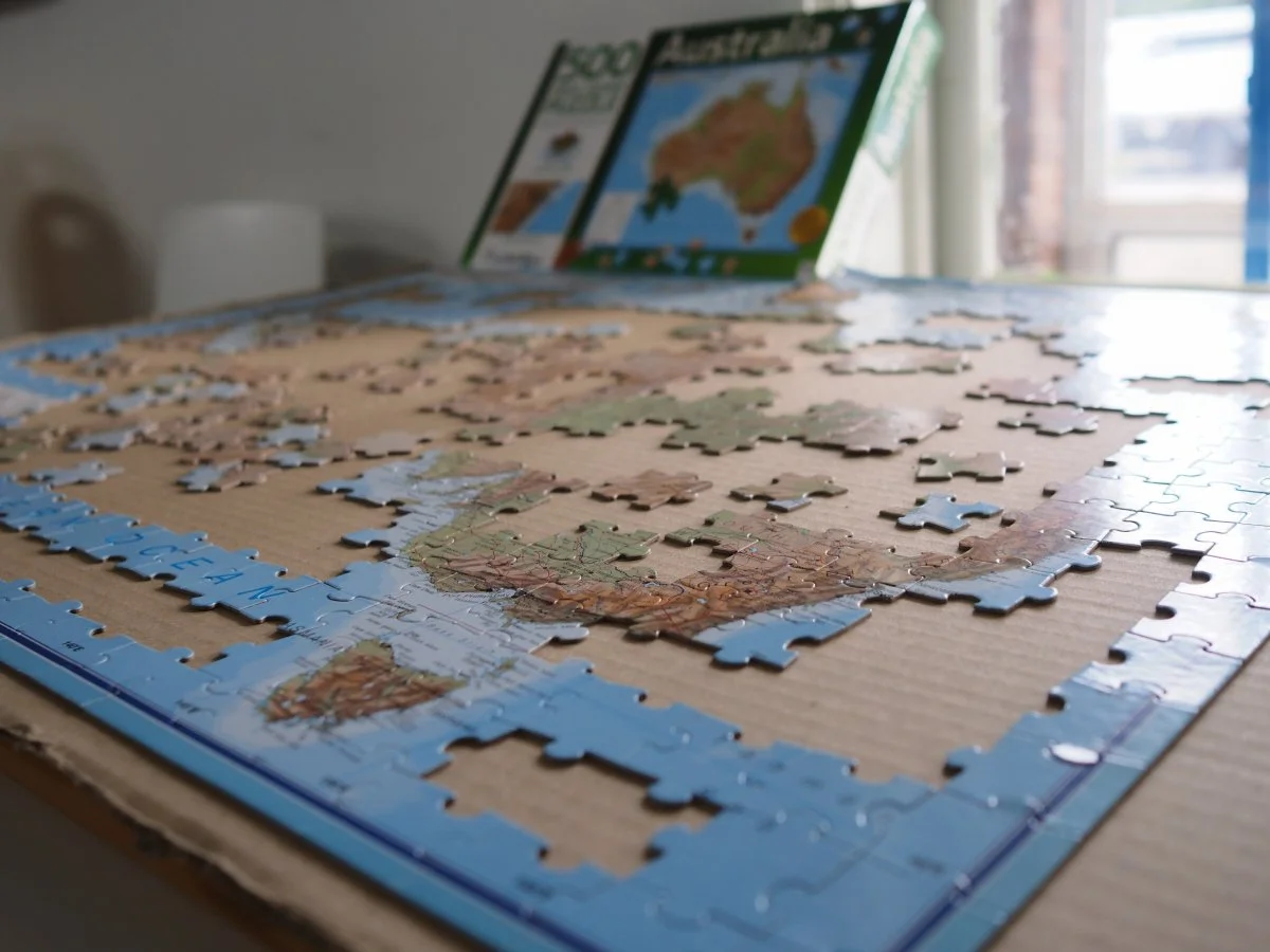 A partially completed jigsaw puzzle of Australia map on a cardboard surface with puzzle boxes in the background.