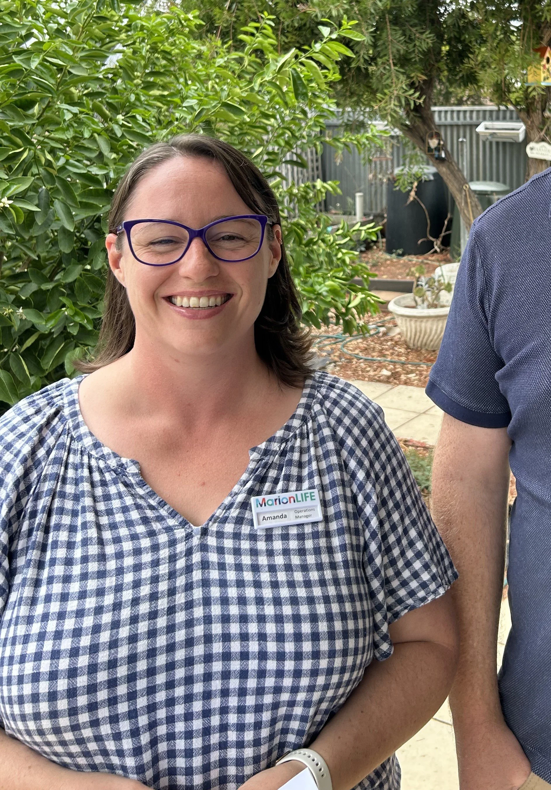 A woman smiling outdoors, wearing glasses and a blue and white checkered dress with a MarionLIFE name tag that reads "Amanda, Operations Manager." She stands in a garden with green foliage, trees, and garden decorations in the background.