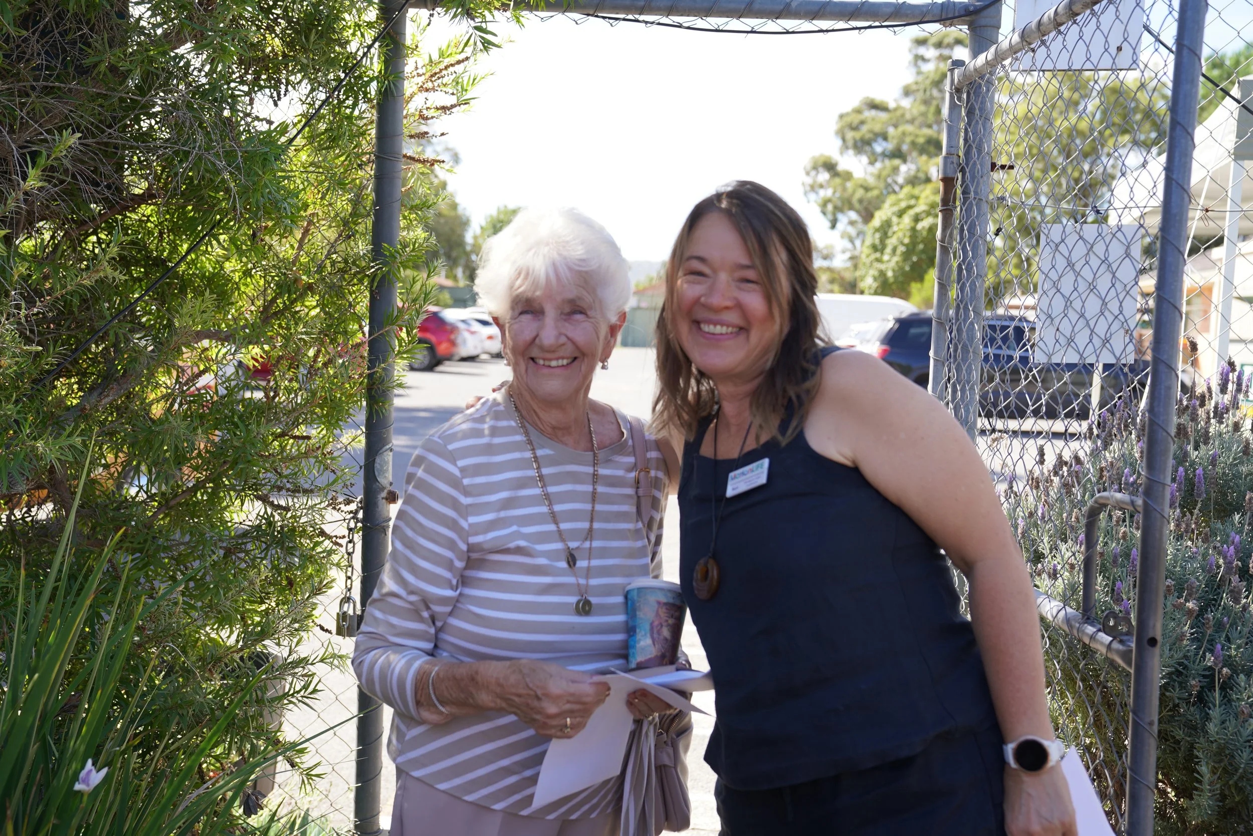 Two women smiling, one older woman with white hair in a striped top holding papers, and a younger woman with dark hair in a black dress standing outside near a fence, greenery, and parked cars.