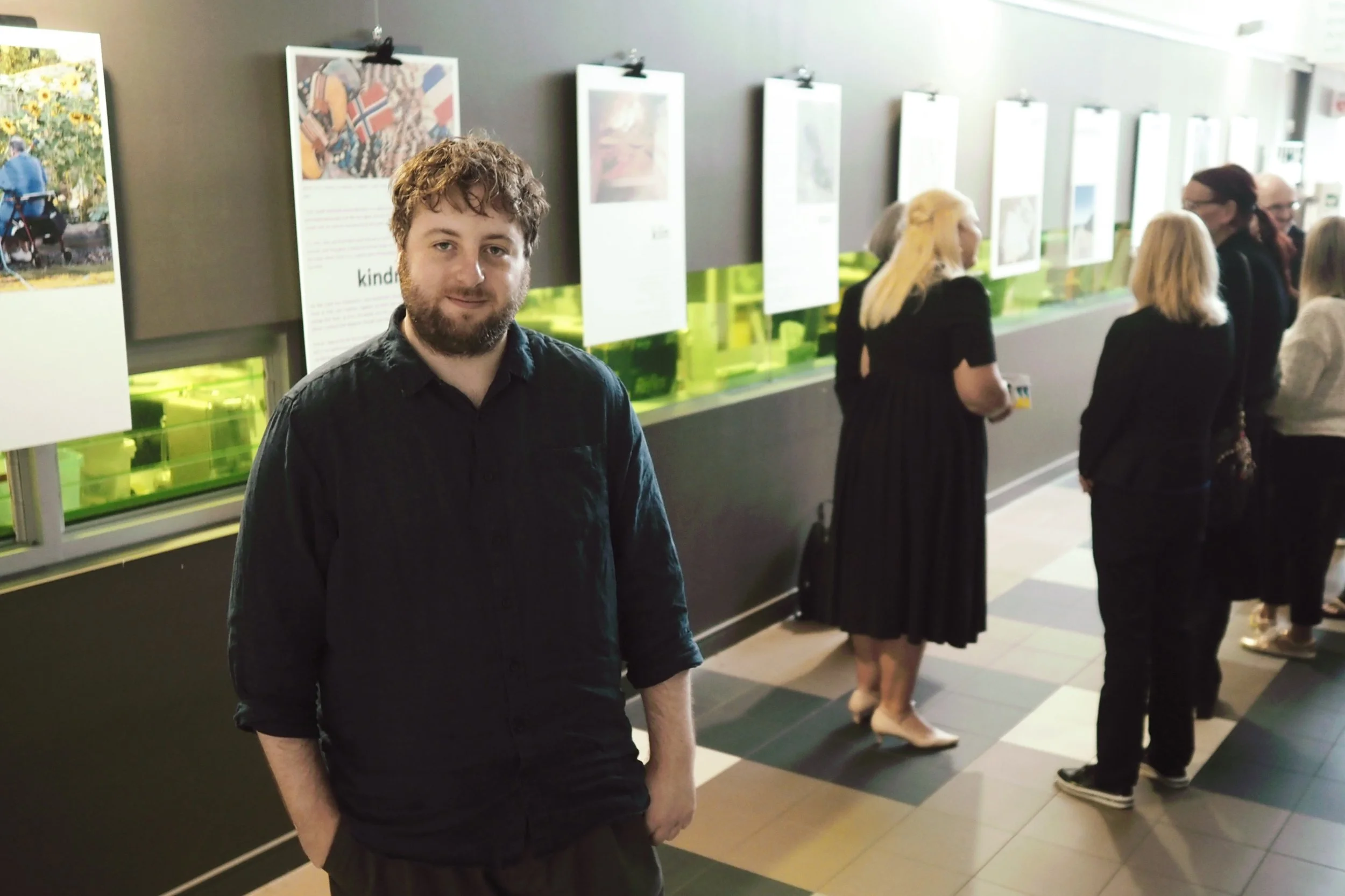 A man with curly hair and a beard wearing a black shirt, standing in an art gallery with people in the background looking at artwork.