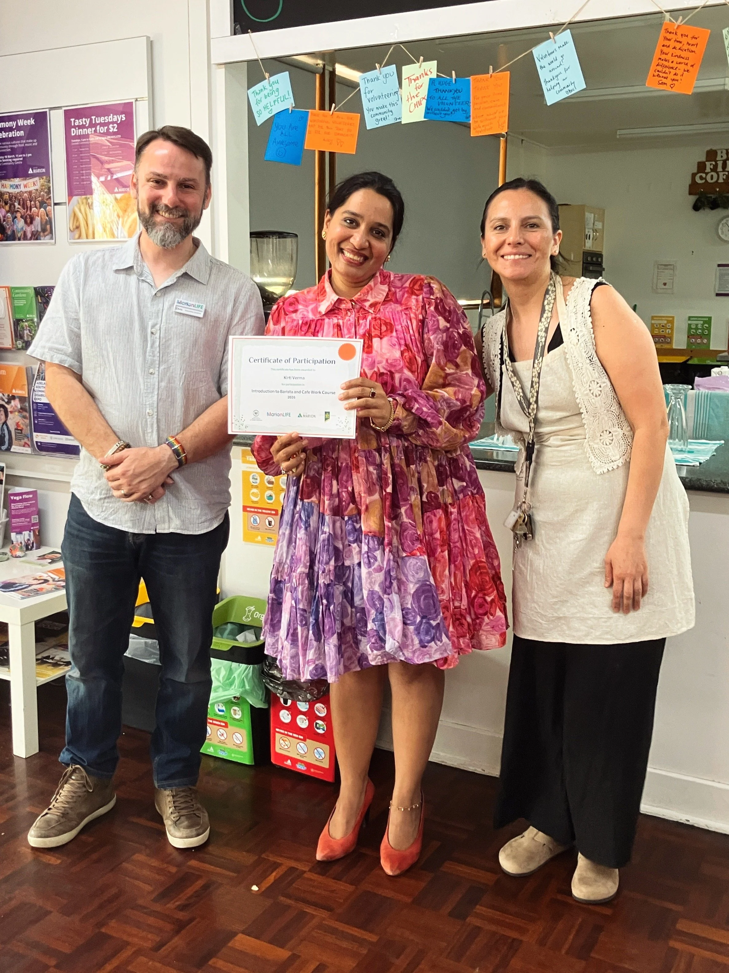 Three people standing close together indoors, smiling at the camera. The person on the left is a man with dark hair and a beard, wearing a light gray shirt and jeans. The person in the middle is a woman with dark hair, wearing a colorful pink and purple dress, holding a certificate of participation. The person on the right is a woman with dark hair pulled back, wearing a sleeveless cream-colored dress with lace details. There are colorful notes on string hanging behind them, celebratory posters on the wall, and a table with informational brochures.
