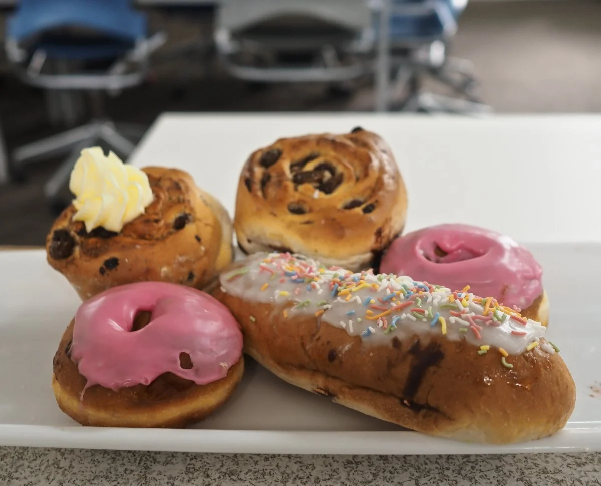 Assorted baked goods on a white platter, including donuts with pink icing and sprinkles, cinnamon rolls, and a pastry with white icing and colorful sprinkles.