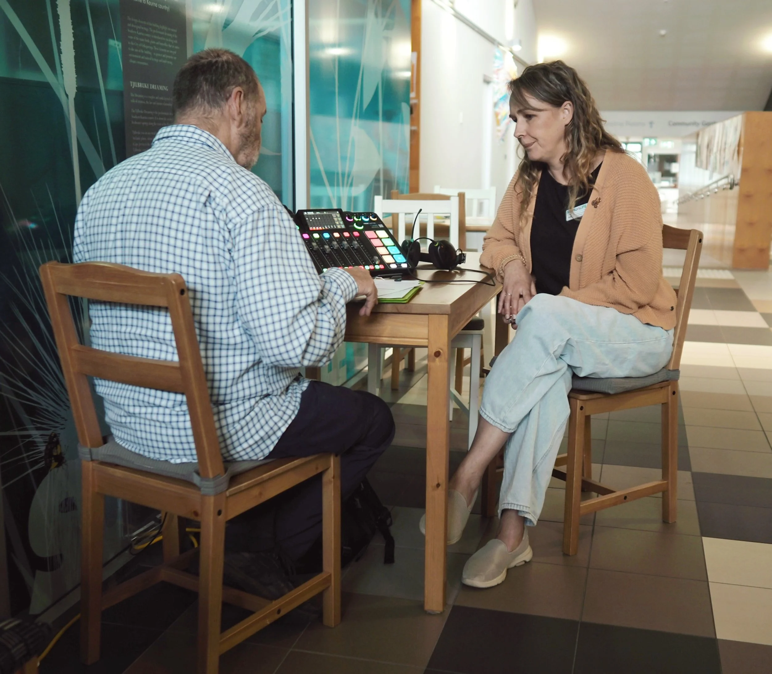 A man and a woman are sitting across from each other at a wooden table in a public indoor space. The man is facing away from the camera, wearing a checkered shirt, interacting with a digital music controller. The woman, dressed in a tan cardigan and black shirt, is sitting with her legs crossed and looking at the man, smiling gently. The background shows a modern, well-lit interior with colorful wall art and glass windows.