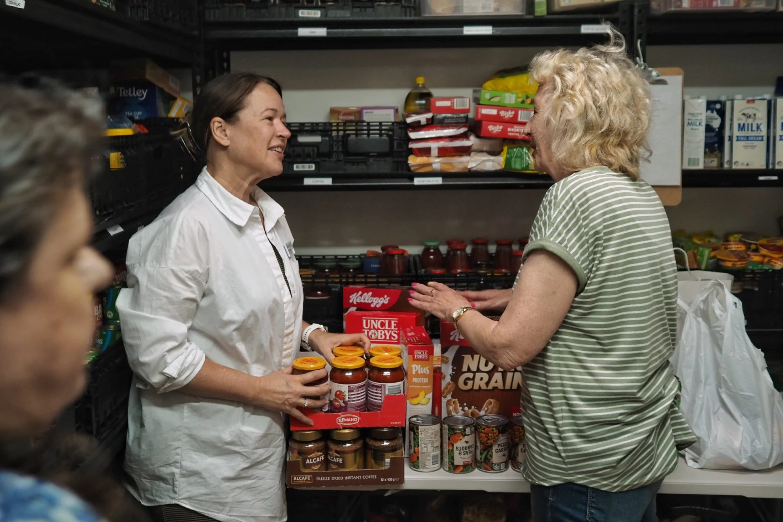 Two women are talking in a grocery store aisle, with the woman on the left holding a jar of sauce and smiling, while the woman on the right is gesturing with her hands. Various food products, including jars, boxes, and canned goods, are visible on the shelves behind them.