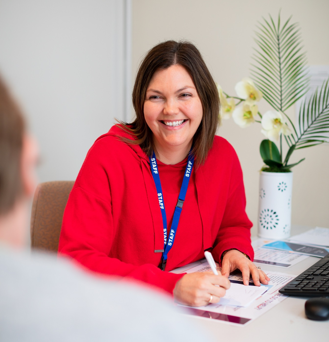 A woman in a red shirt with a lanyard sitting at a desk, smiling and writing notes, with decorative flowers in a vase nearby.