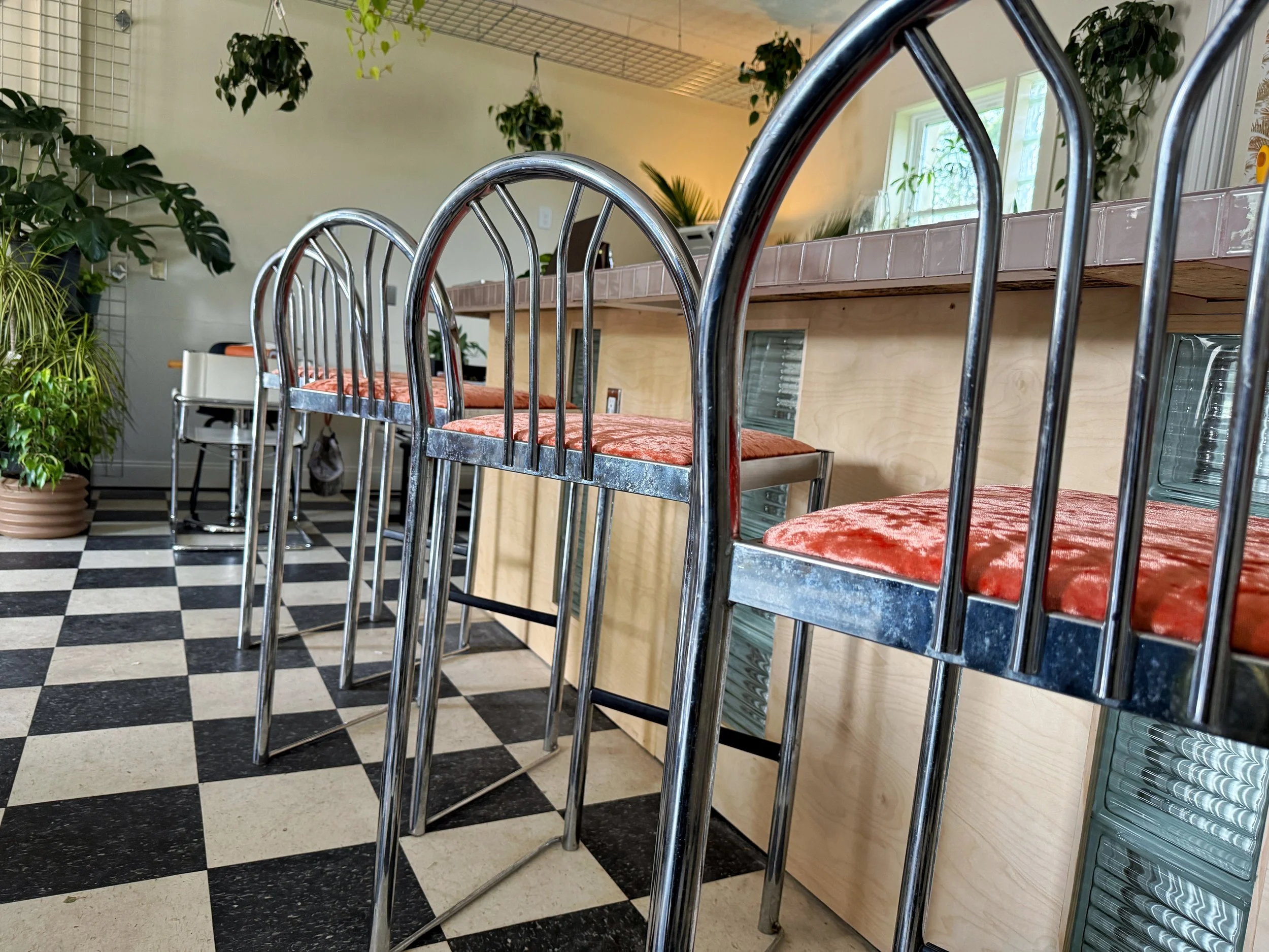 A row of metal barstools with pink velvet cushions in a cafe or restaurant, with large green plants and a black-and-white checkered floor.