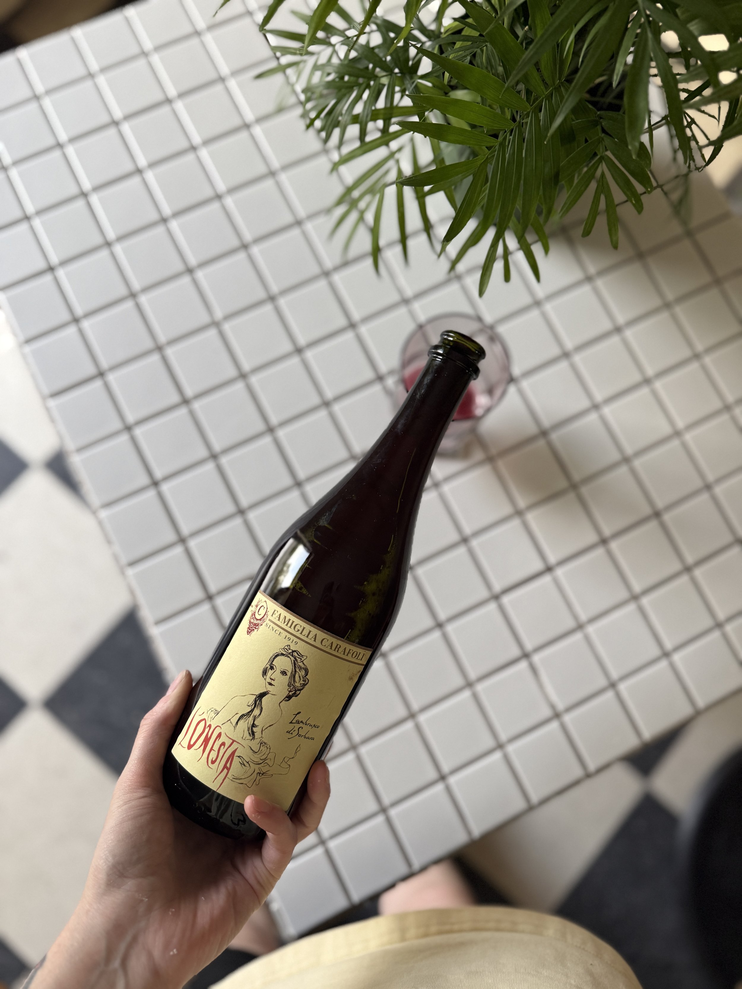 A person holding a bottle of wine with a beige label featuring an illustration of a woman, set against a background of white tile flooring and green plants.