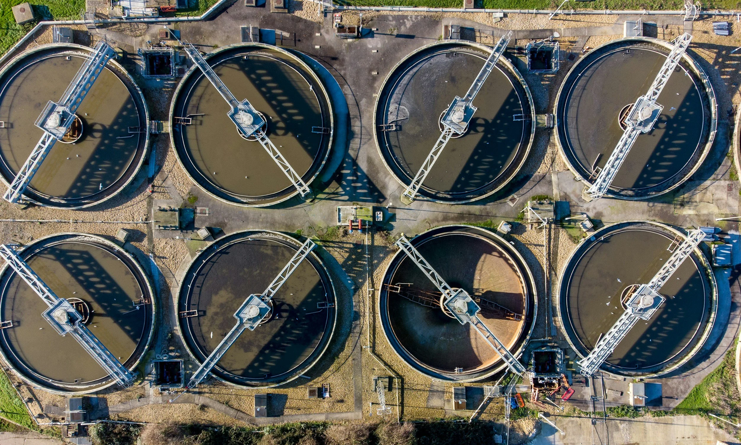 Aerial view of an industrial wastewater treatment plant with eight circular clarifiers, each equipped with mechanical arms and covered with shadows from the structures.
