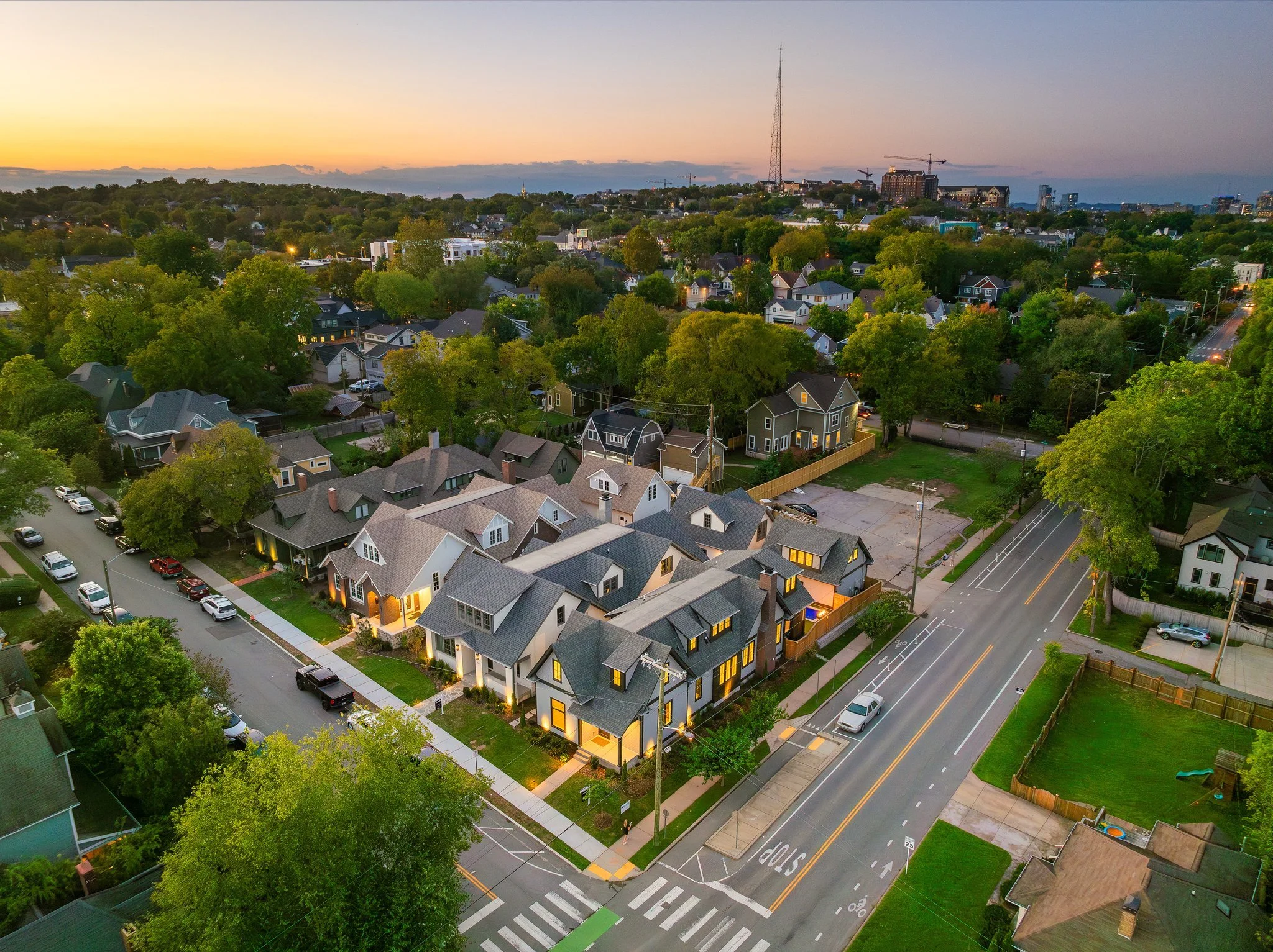 Aerial view of a residential neighborhood at sunset, showing houses, trees, streets, parked cars, and a park with a few people walking.