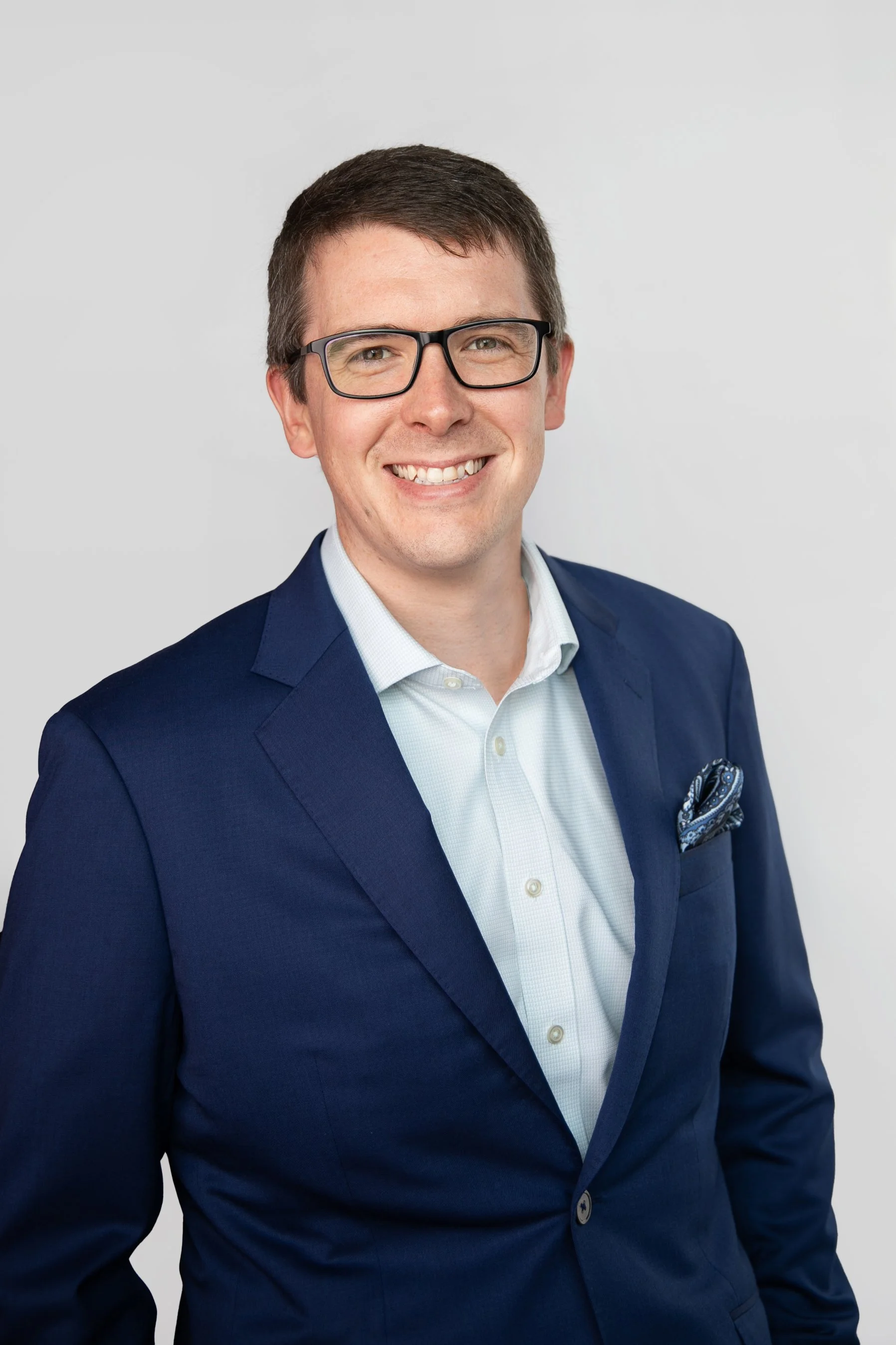 Portrait of a young man with short brown hair, glasses, and a smile, wearing a navy blue suit jacket, a white shirt, and a patterned pocket square, against a plain grey background.