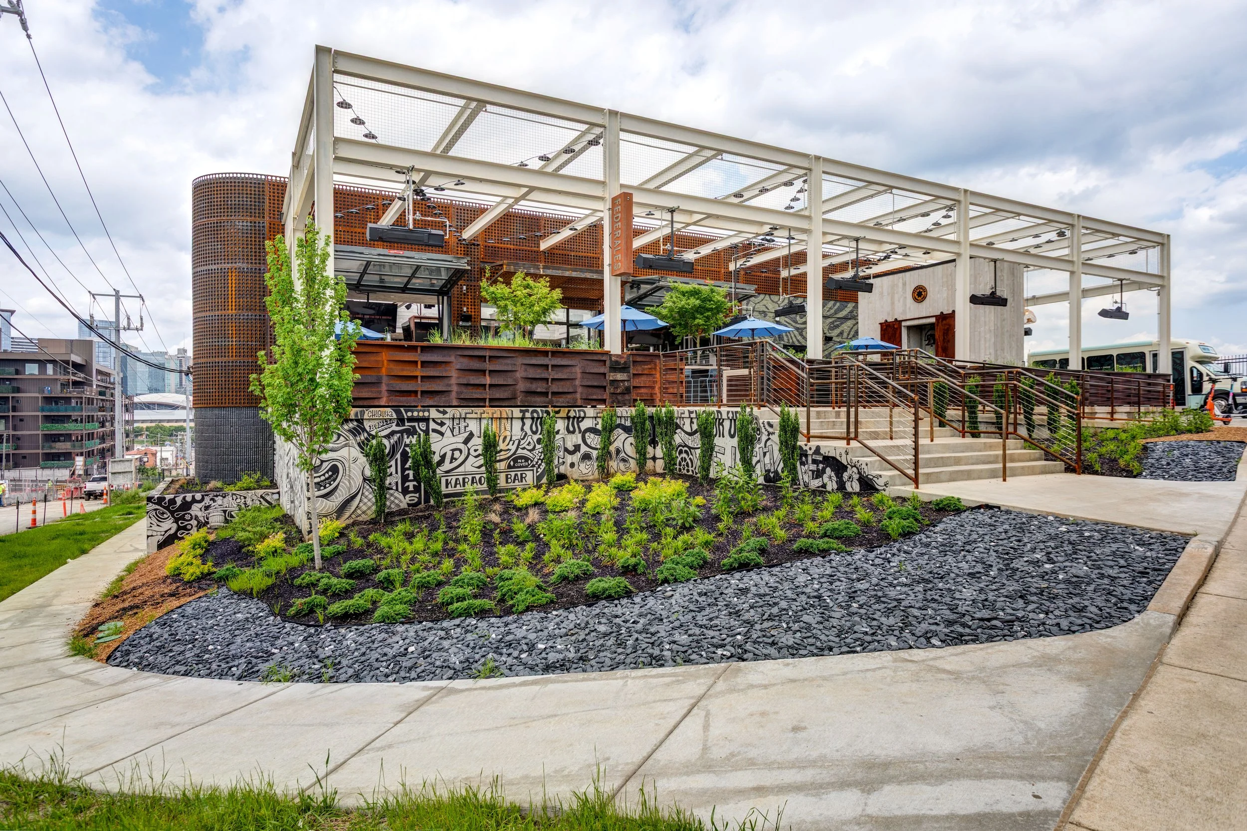 Outdoor rooftop bar and dining area with tables, chairs, umbrellas, and greenery, located on an elevated terrace with stairs and a decorative mural on the wall.