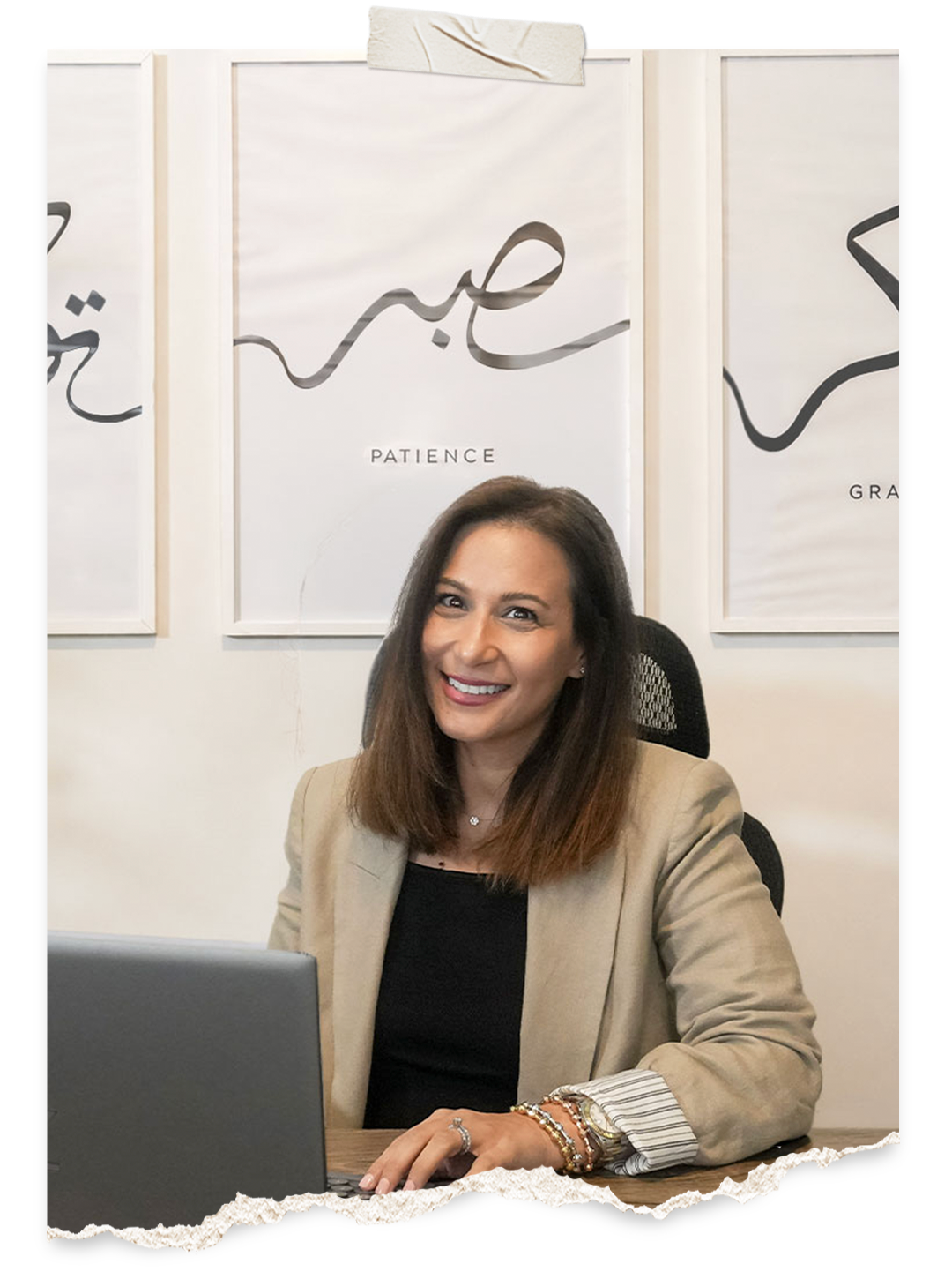 A woman with brown hair, wearing a beige blazer and black top, sitting at a desk with a laptop, smiling at the camera. Behind her are framed artworks with words 'Patience' and other Arabic script.
