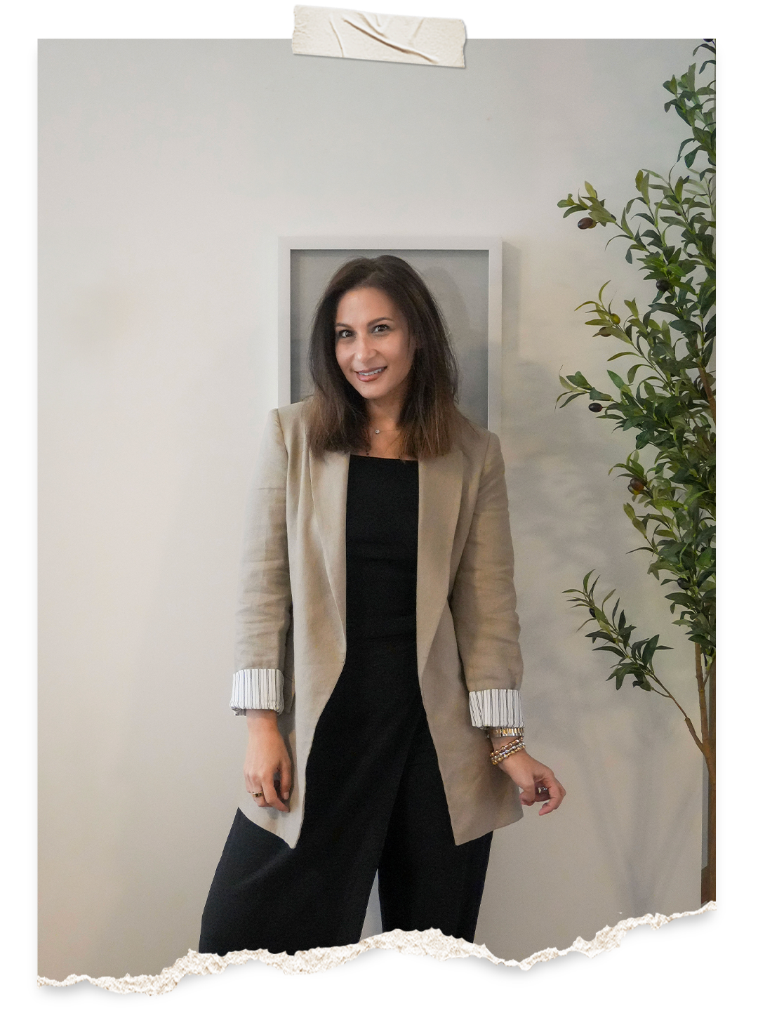 A woman with shoulder-length brown hair smiling at the camera, wearing a beige blazer with striped cuffs over a black outfit, standing indoors next to a green leafy plant, with a torn paper effect border at the bottom of the image.