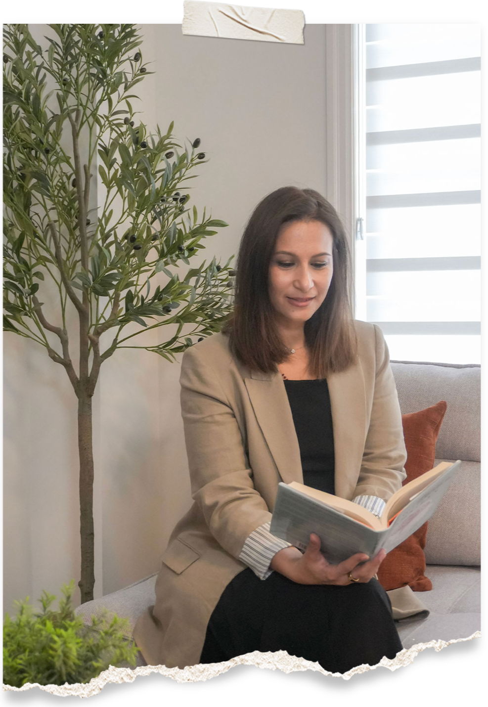 A woman with shoulder-length brown hair, wearing a beige blazer and a black top, sitting on a light-colored couch with a brown pillow, reading a book. There is a potted plant with green leaves and black berries next to her, and a window with blinds in the background.