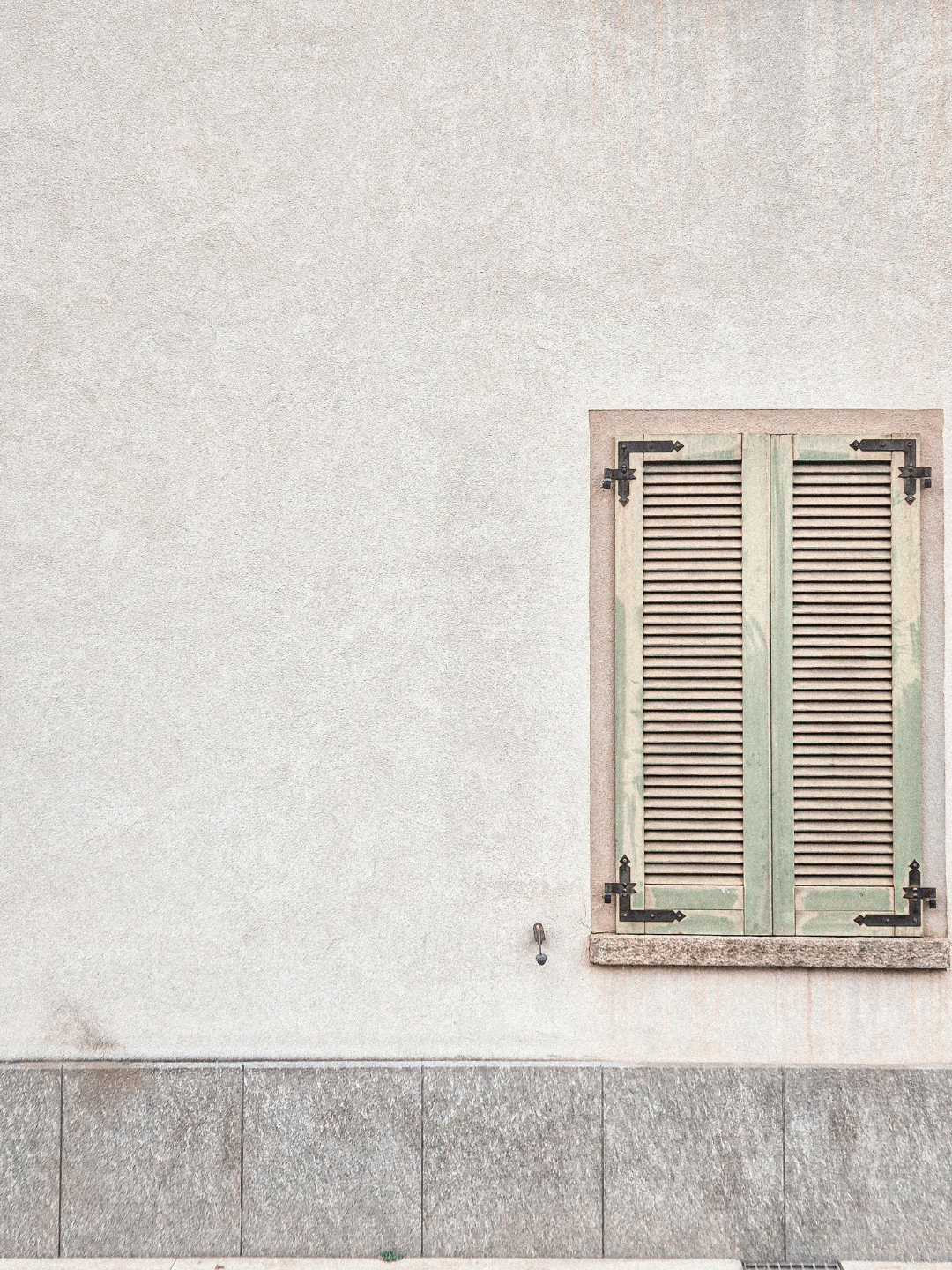 A weathered wooden window with green shutters and black metal hinges on a plain white exterior wall with a concrete base.
