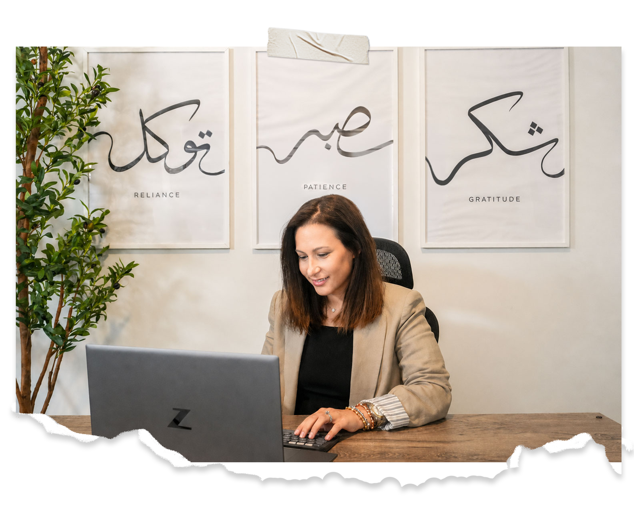 A woman sitting at a wooden desk using a laptop, with a wall behind her decorated with three framed words: 'Reliance,' 'Patience,' and 'Gratitude.' There is a green plant on the left side of the image.