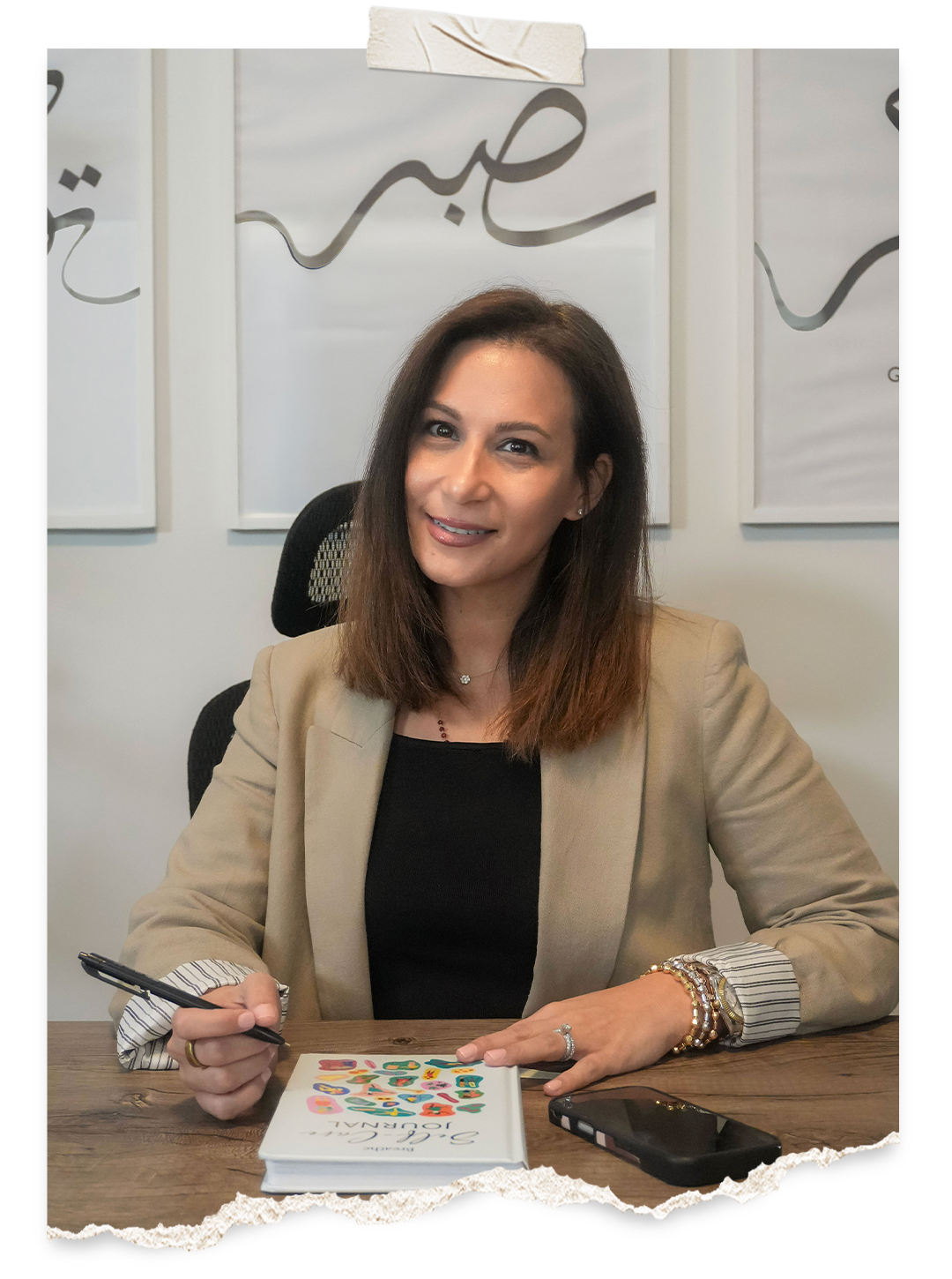 A woman with shoulder-length brown hair sitting at a wooden table, holding a black pen, with a book titled 'Soulful Journaling' and a smartphone on the table. Behind her are white wall decorations with cursive writing.