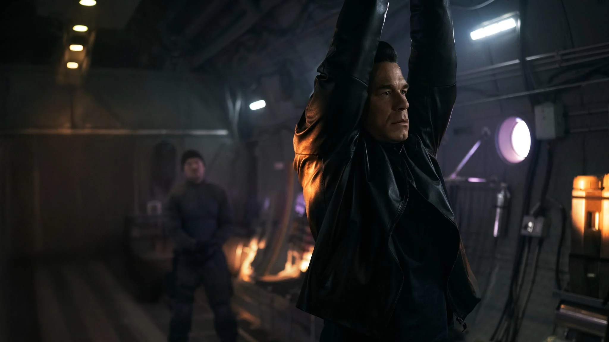 A man wearing a black leather jacket with his hands overhead, in a dark industrial or sci-fi setting with dim lighting and blurred background figures.