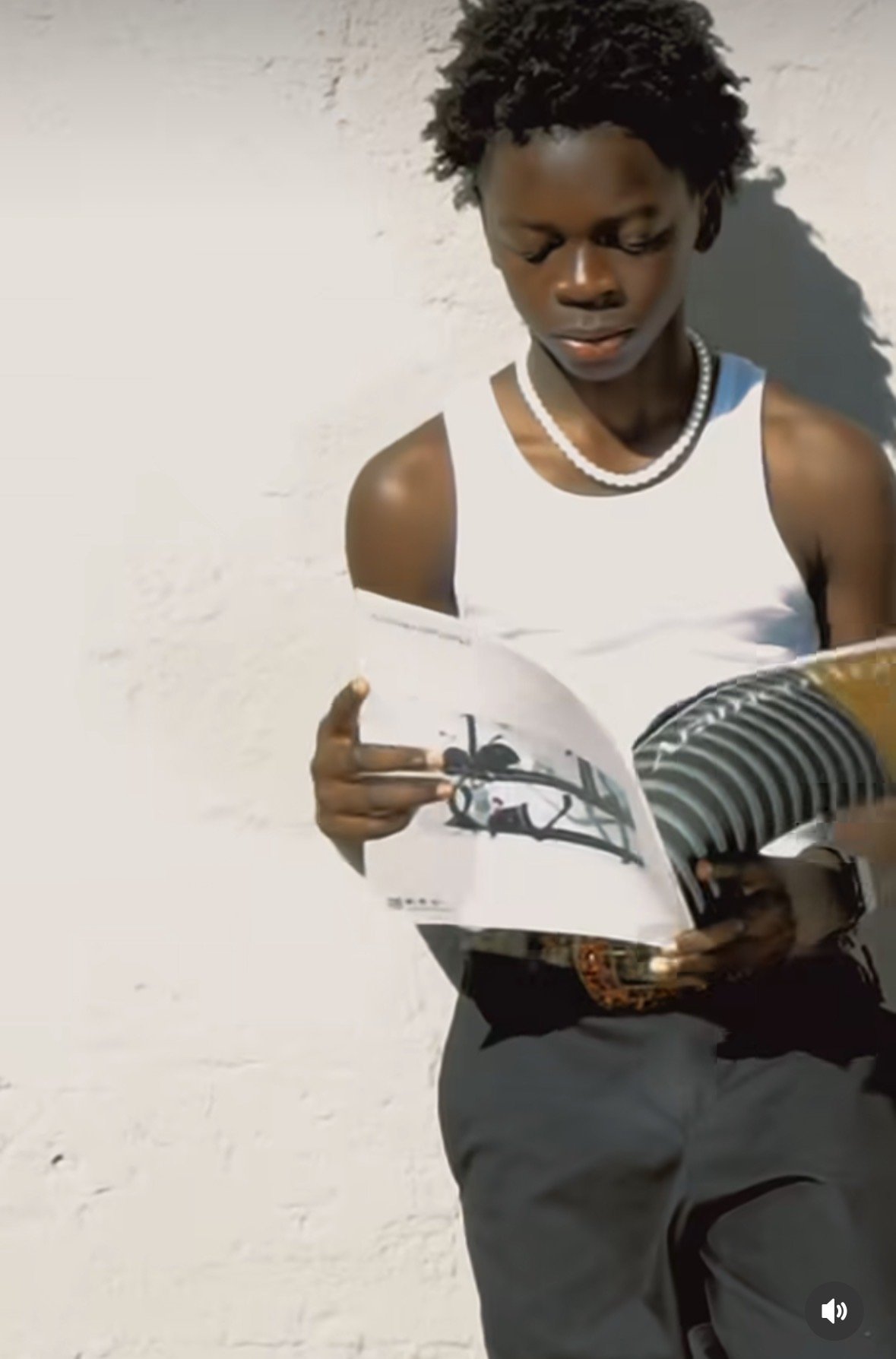 A person reading a newspaper outdoors against a white wall, with the sun casting a shadow.
