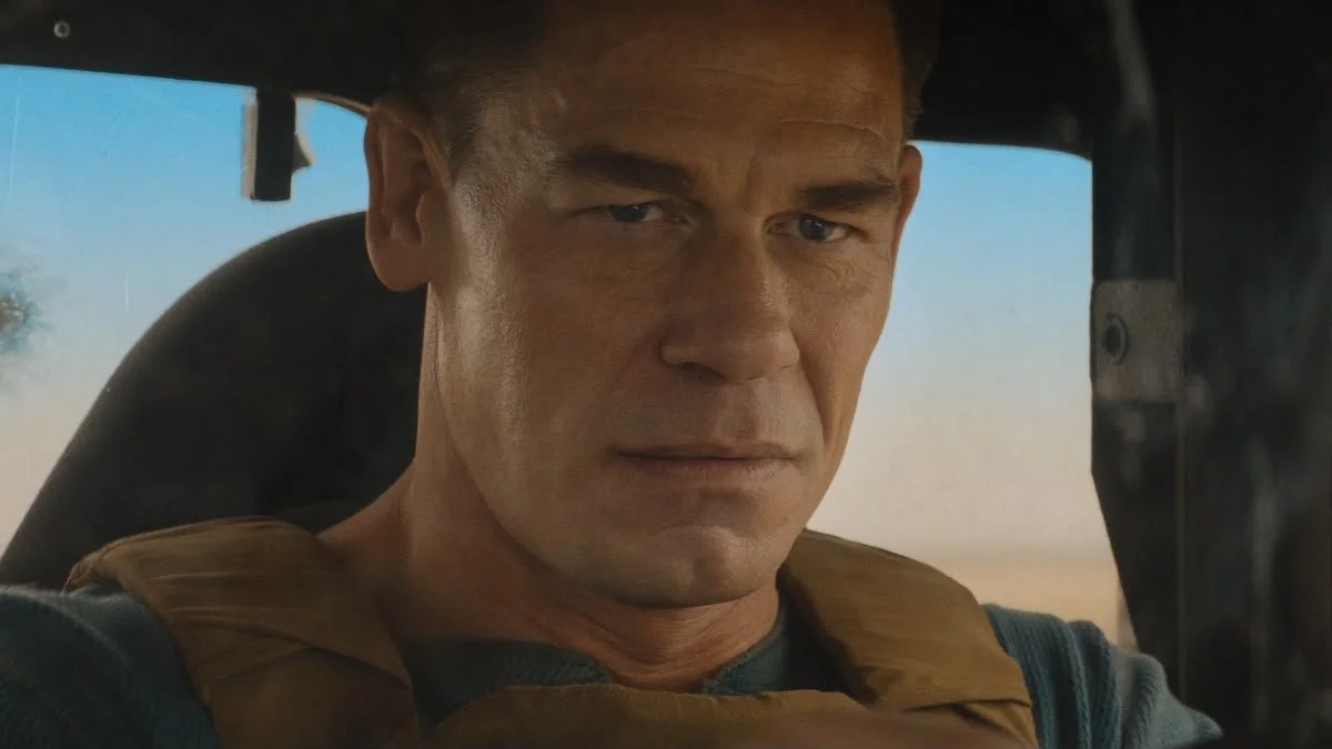 Close-up of a man with short hair and a serious expression, driving a vehicle in a desert landscape.