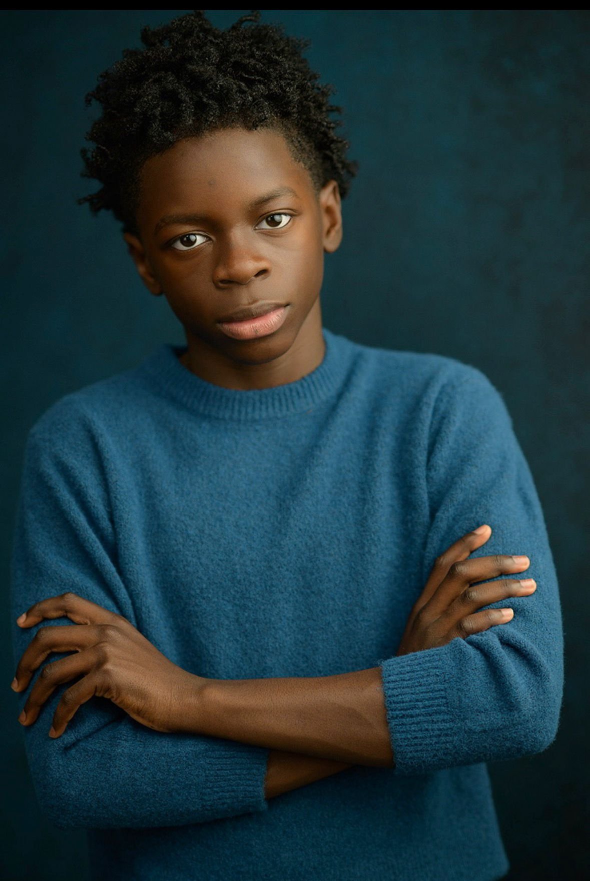 Portrait of a young person with dark skin, curly hair, wearing a blue sweater, crossing arms, against a dark blue background.