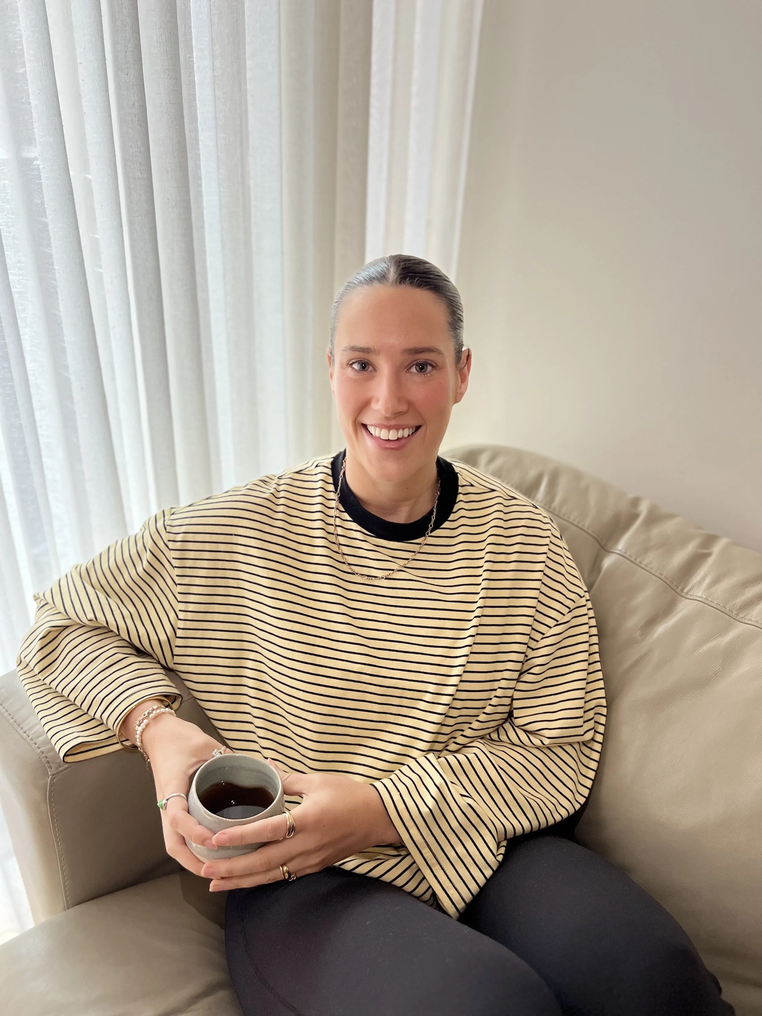 Young woman with dark hair in a bun, smiling, sitting on a beige couch, holding a gray mug of coffee, wearing a striped beige and black shirt, jewelry, in a room with white curtains.