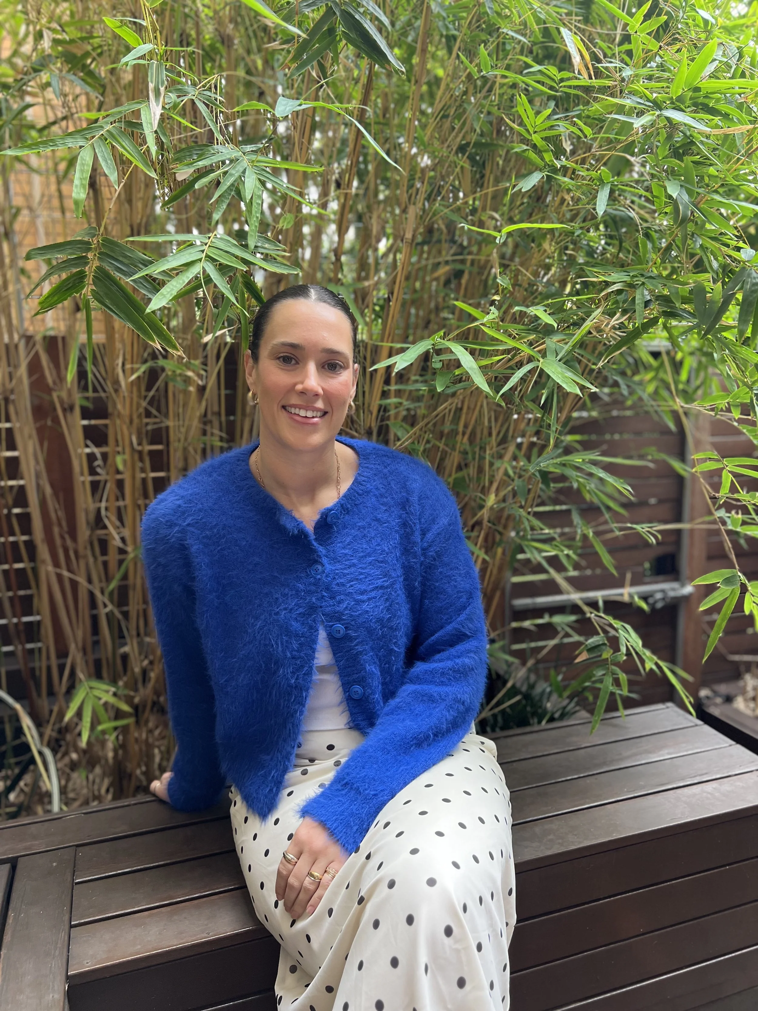 A woman sitting on a wooden bench in a garden with green bamboo plants in the background, wearing a bright blue sweater and a white polka dot skirt.