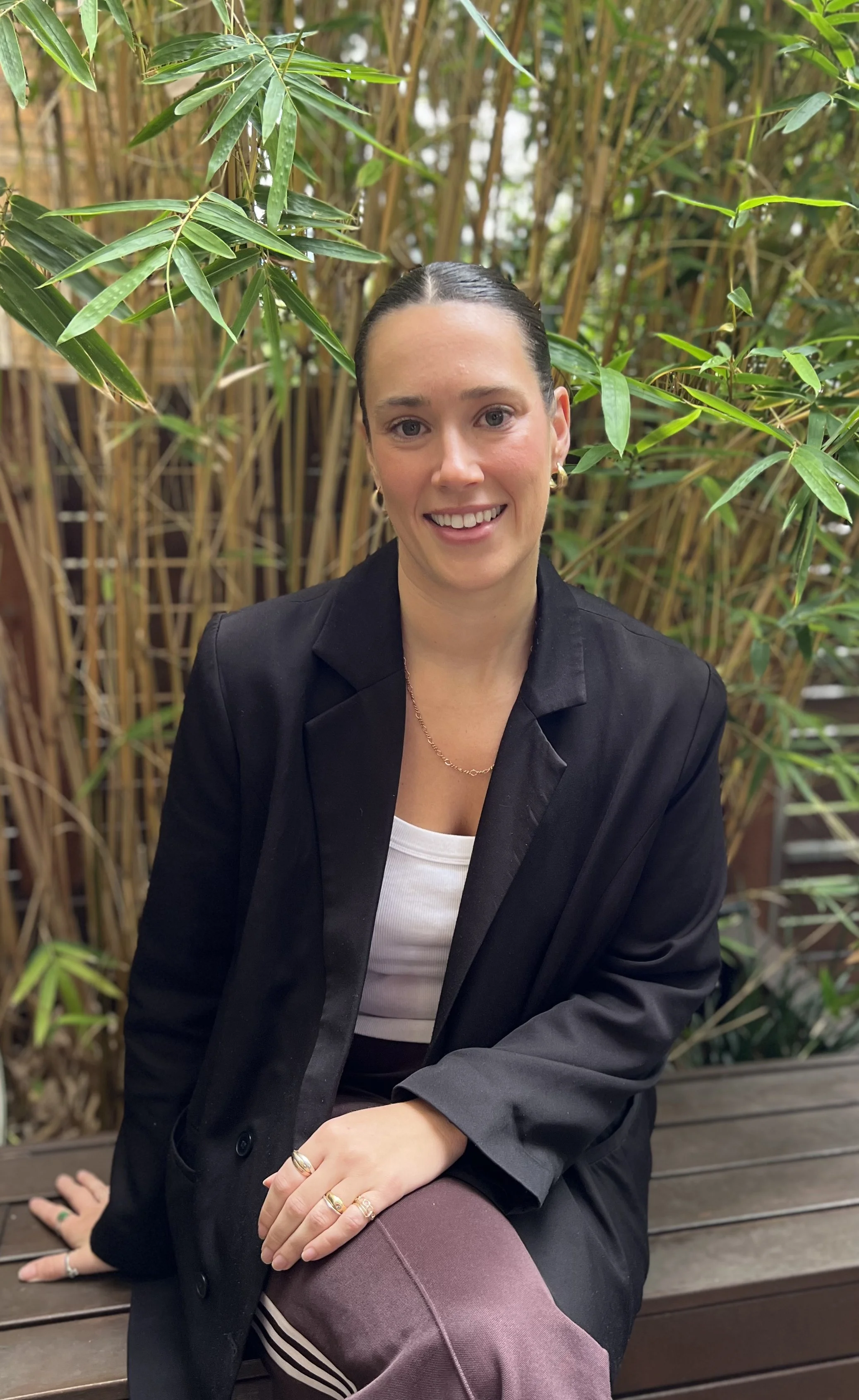 A woman with dark brown hair pulled back, wearing a black blazer, white shirt, and purple pants, sitting on a wooden bench outdoors surrounded by green bamboo plants, and smiling at the camera.