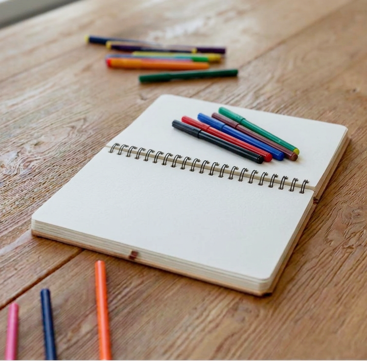 Open blank spiral notebook with markers and colored pencils scattered around on wooden table.