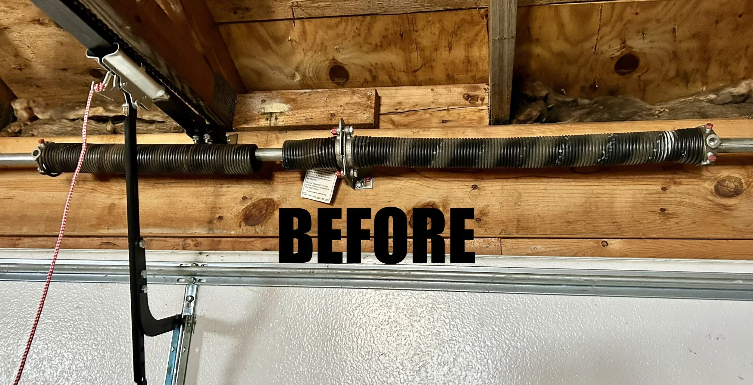 Garage door opener mounted on a wooden garage ceiling with black coiled safety cable and metal brackets, with the word 'BEFORE' written in black text at the bottom.