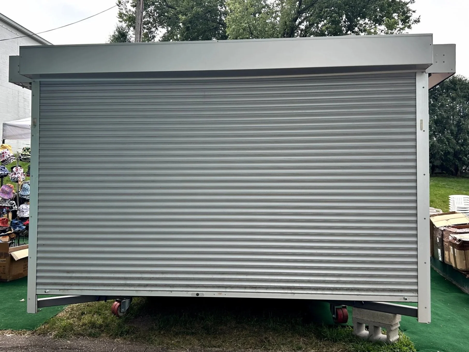 A closed metal rolling shutter on a small structure, with surrounding trees and a grassy area nearby.