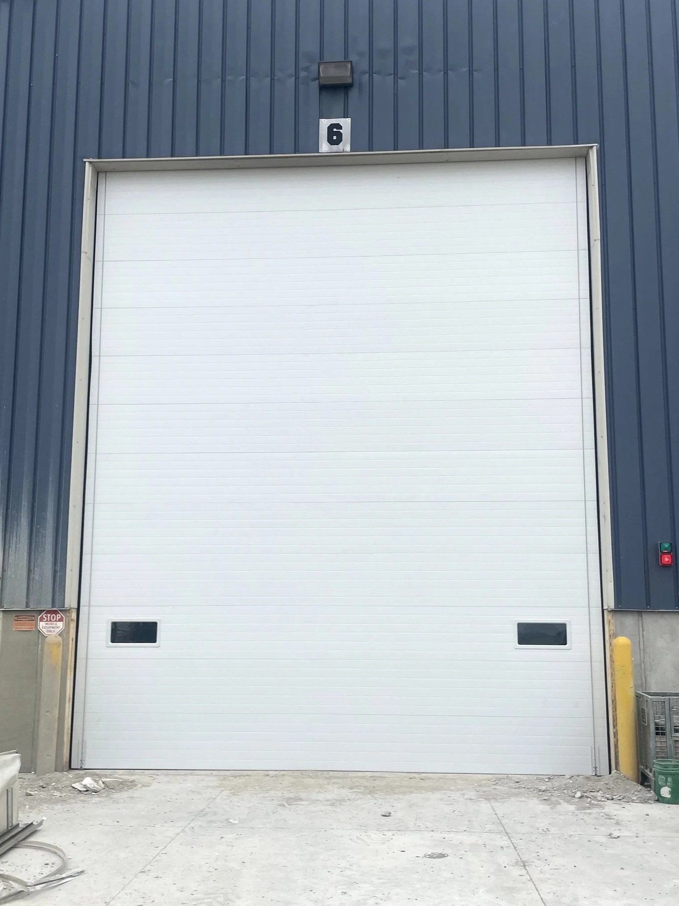 Industrial building with a large, closed white roll-up door, blue metal siding, and a small rectangular window at the bottom. A number 6 sign is mounted above the door. There are yellow safety posts on either side of the door, with a red STOP sign an
